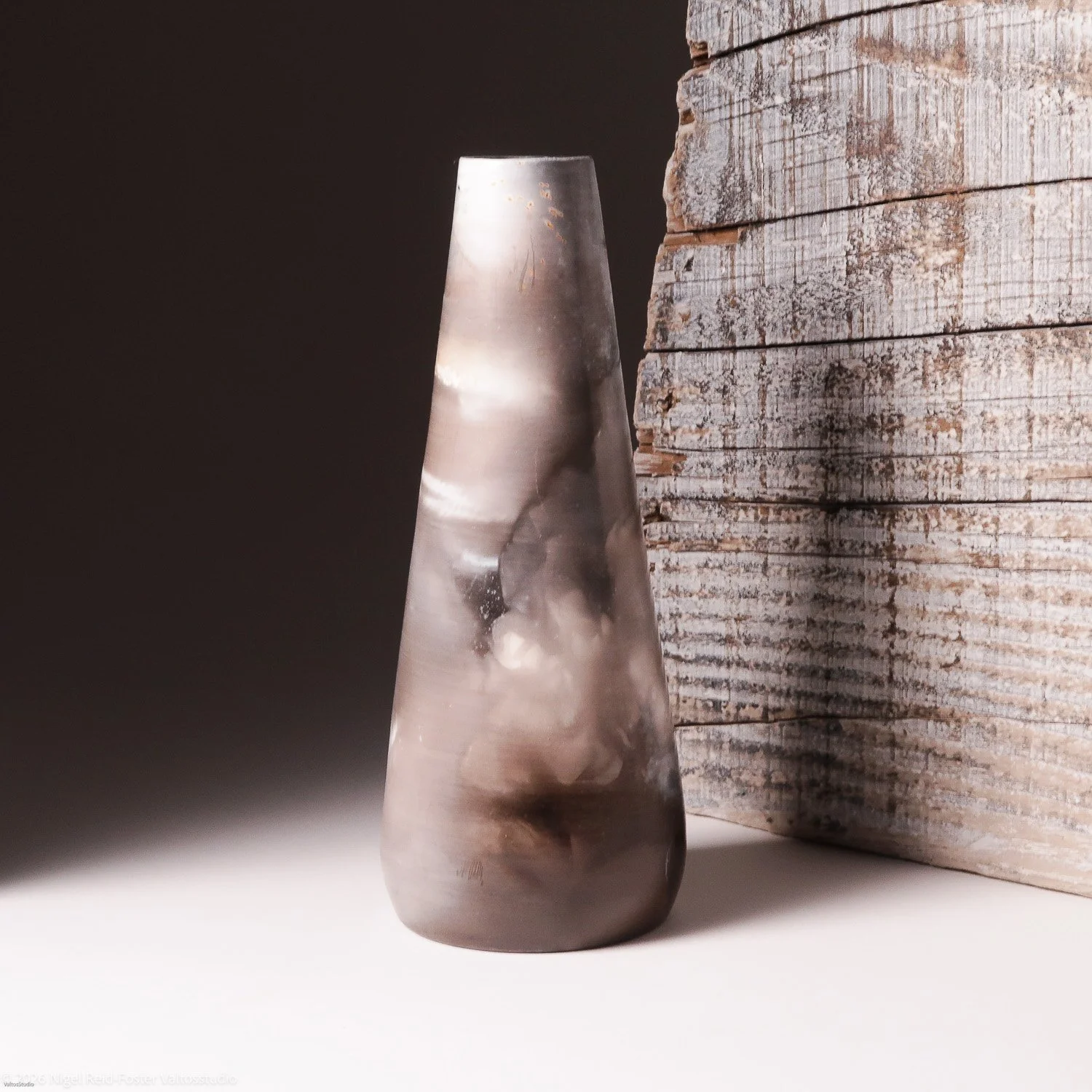 A tall, smooth, ceramic vase with a swirl pattern, placed on a white surface next to a weathered wooden wall.