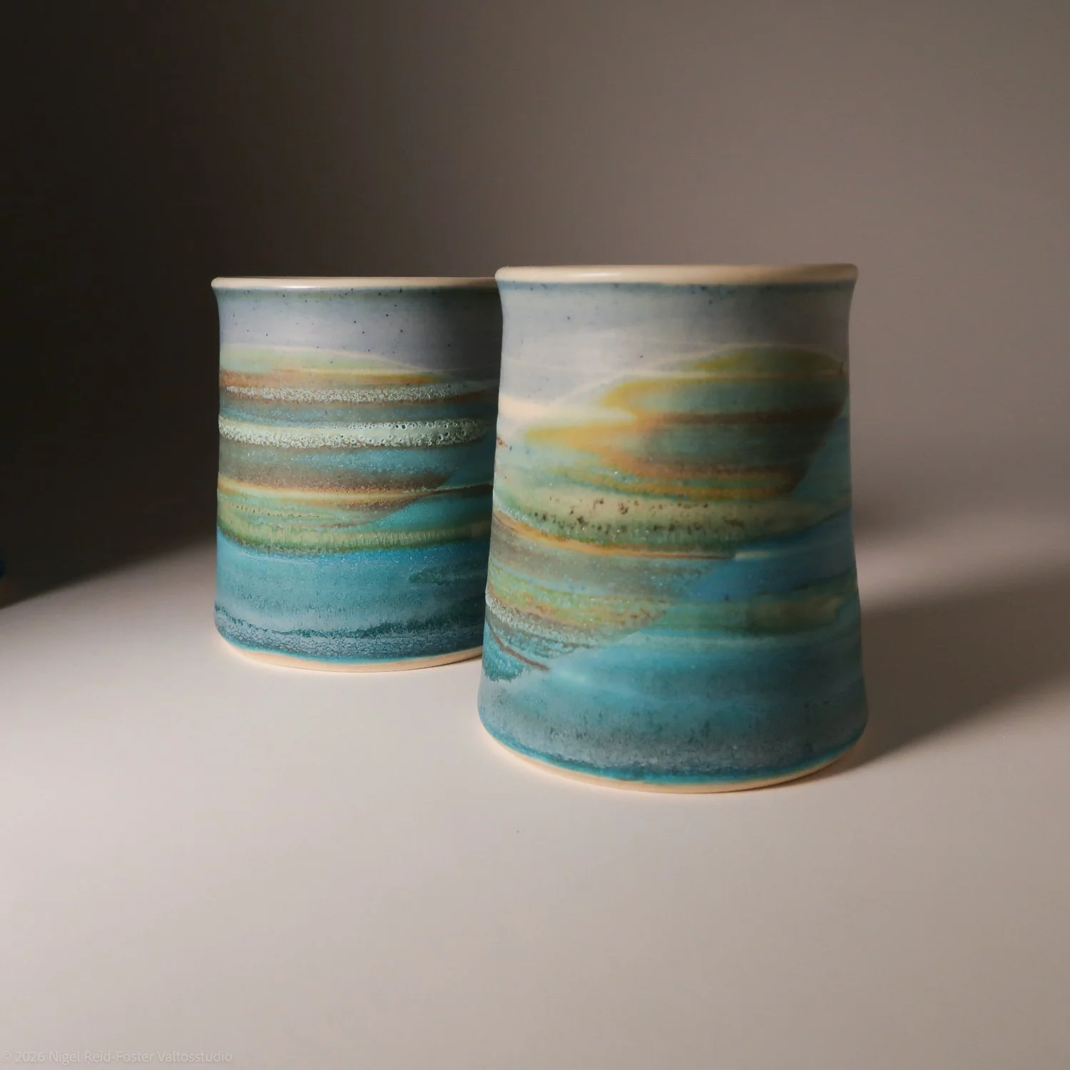 Two ceramic cups with a colorful, swirling marble-like pattern in shades of blue, green, yellow, and brown, placed on a white surface with a dark background.