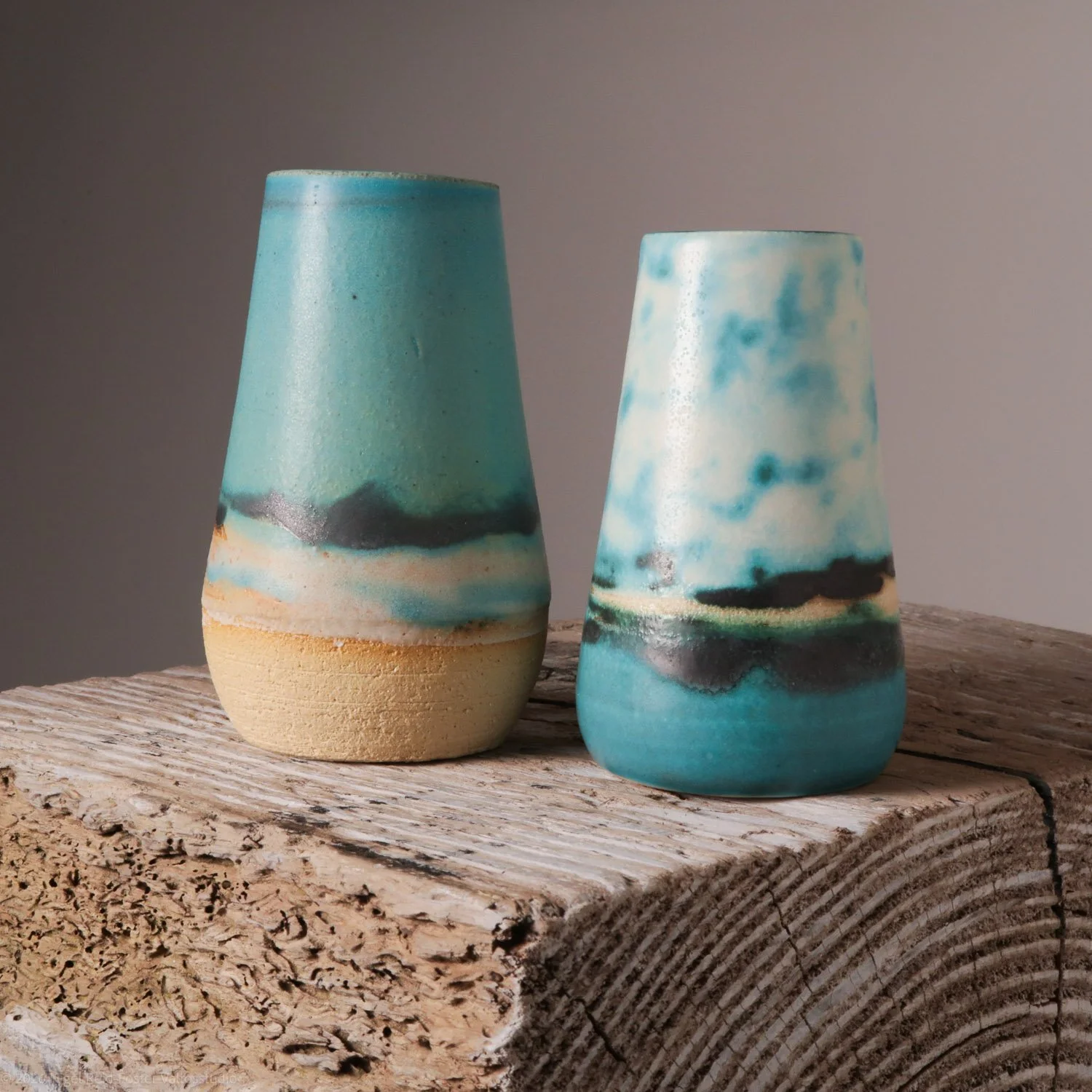 Two ceramic vases with blue, black, and beige abstract glaze, placed on a textured wooden surface.