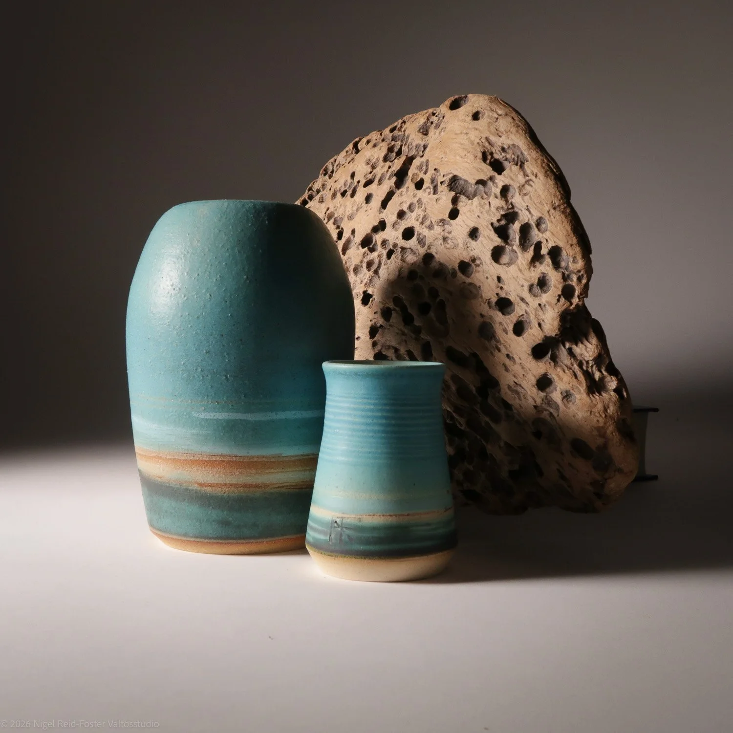 Three pottery vases with varying shades of blue and beige, positioned in front of a large, textured piece of volcanic rock.