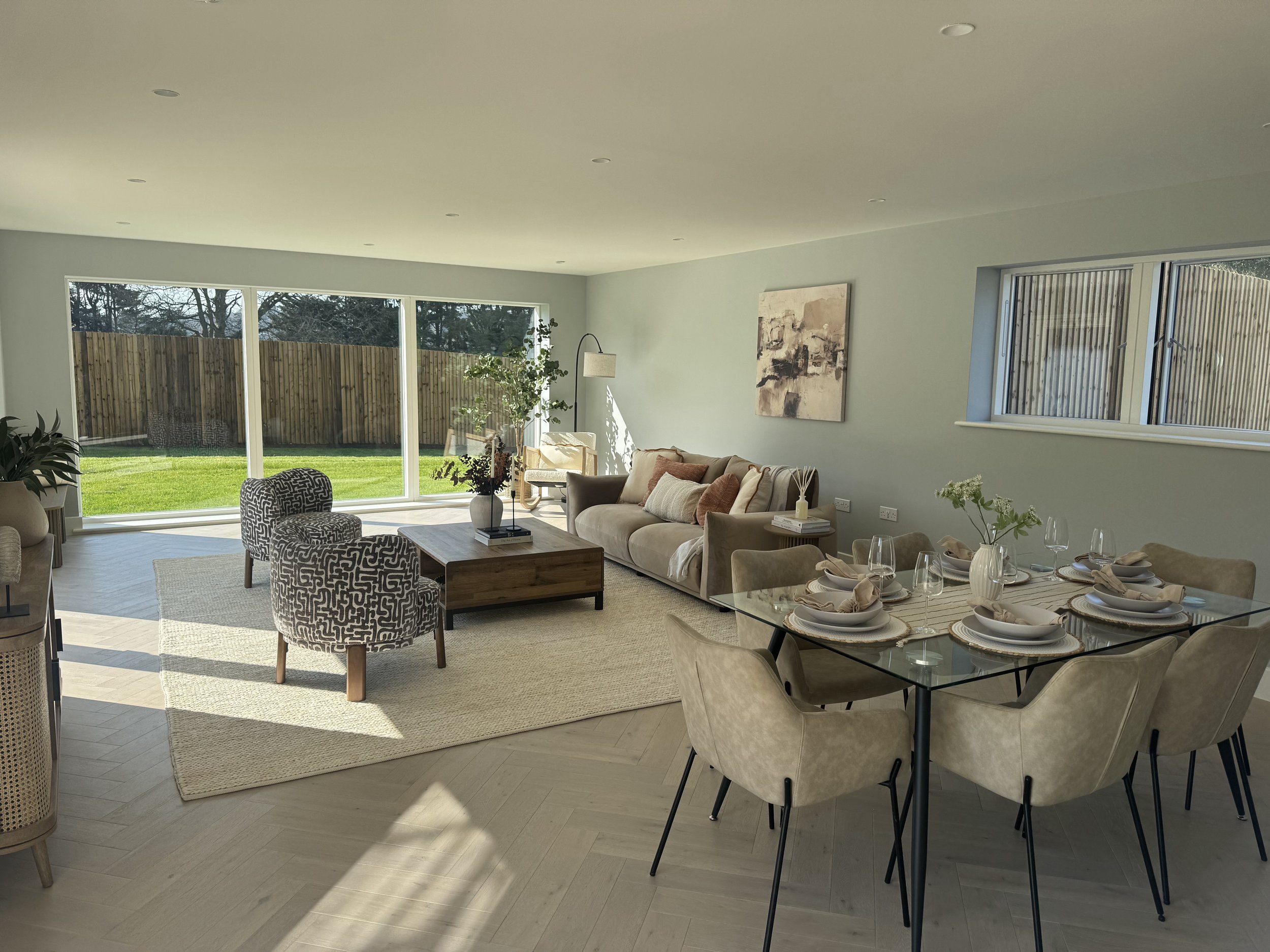 Bright living and dining room with large window and sliding glass door, beige sofa, patterned armchairs, dining table set with plates, glasses, and napkins, and a view of a green backyard.