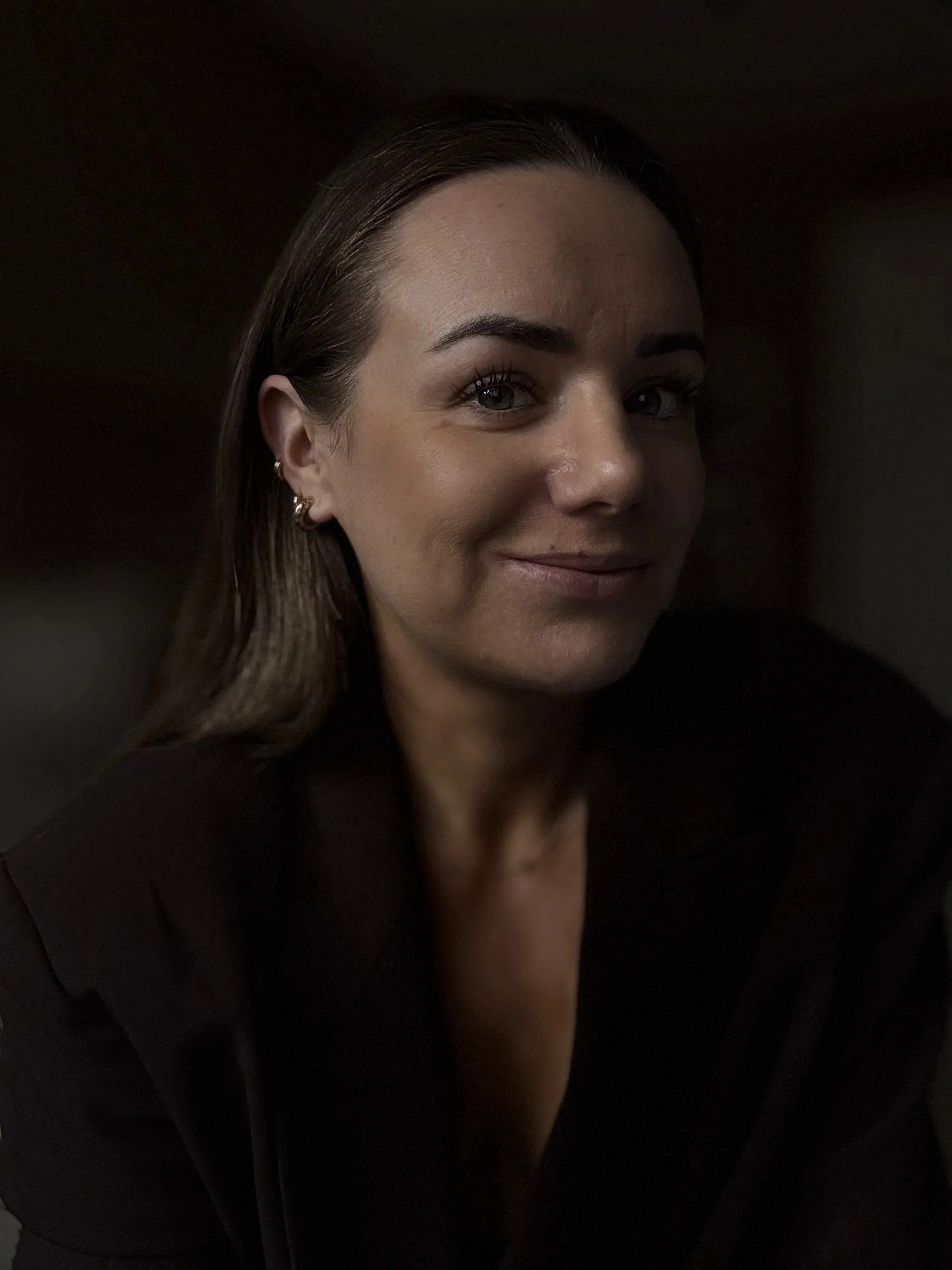 Close-up of a smiling woman with long brown hair, earrings, and a black blazer against a dark background.
