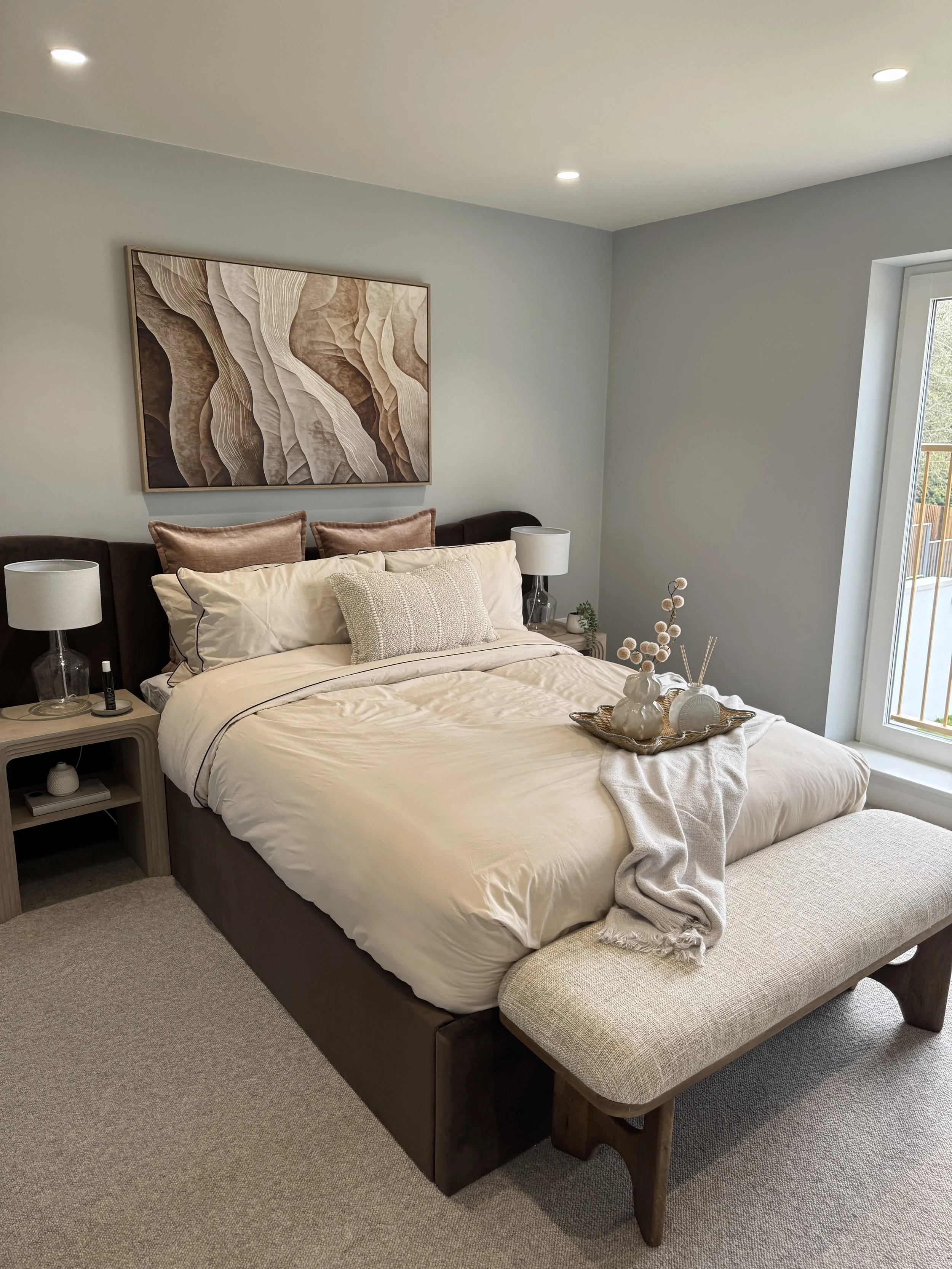 A neatly made bed with beige and brown pillows, a decorative tray with vases and ornaments on the blanket, bedside tables with lamps, a large window, and a modern abstract painting above the bed in a bedroom.