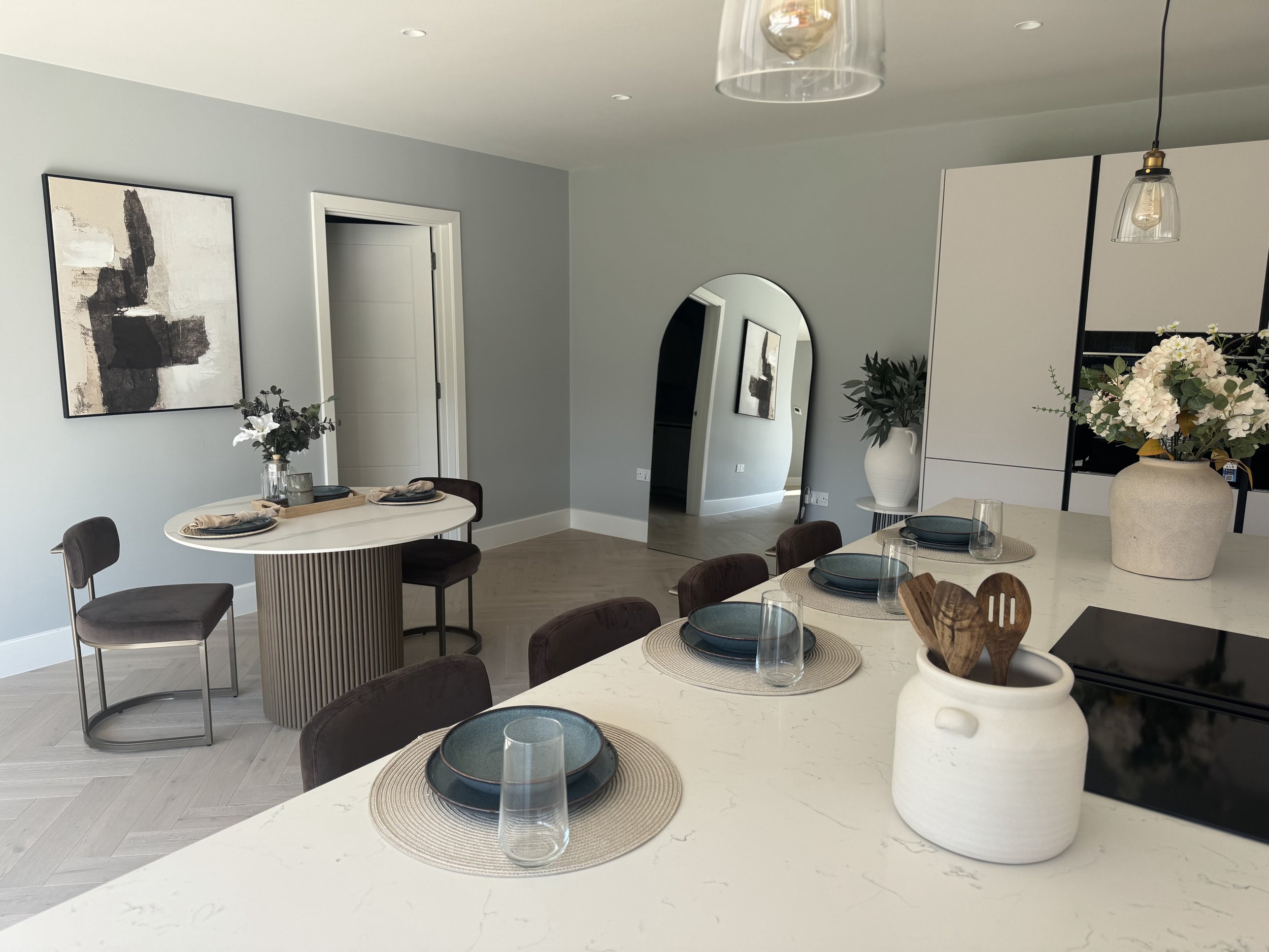 Modern dining area with a white marble table set with blue plates and glasses, surrounded by dark chairs, and decorated with vases of flowers, with a minimalist aesthetic and neutral color palette.