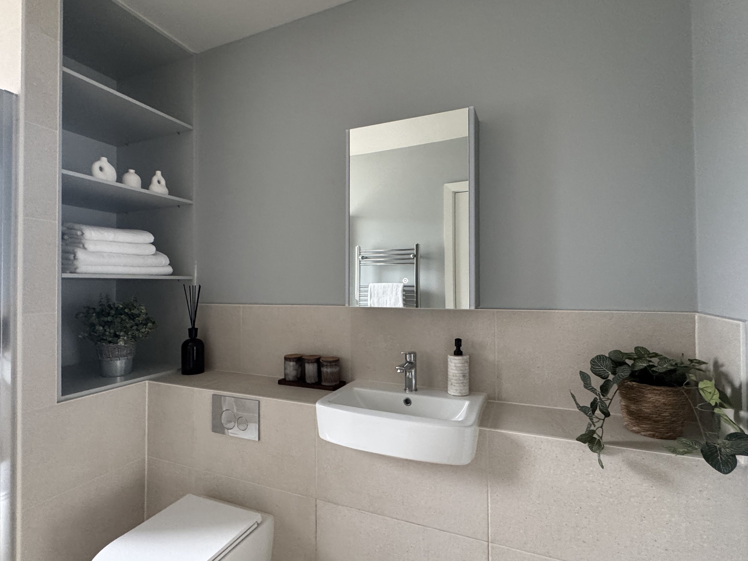 A bathroom with a small white sink, a mirror above, beige tiled wall, open shelves with white towels and decorative items, a potted plant on the right, and a black soap dispenser on the sink.