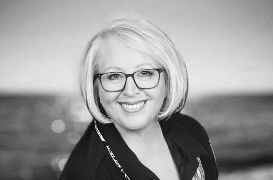 Black and white portrait of a smiling woman with short blonde hair, glasses, and a scarf around her neck, with a blurred beach and ocean background.