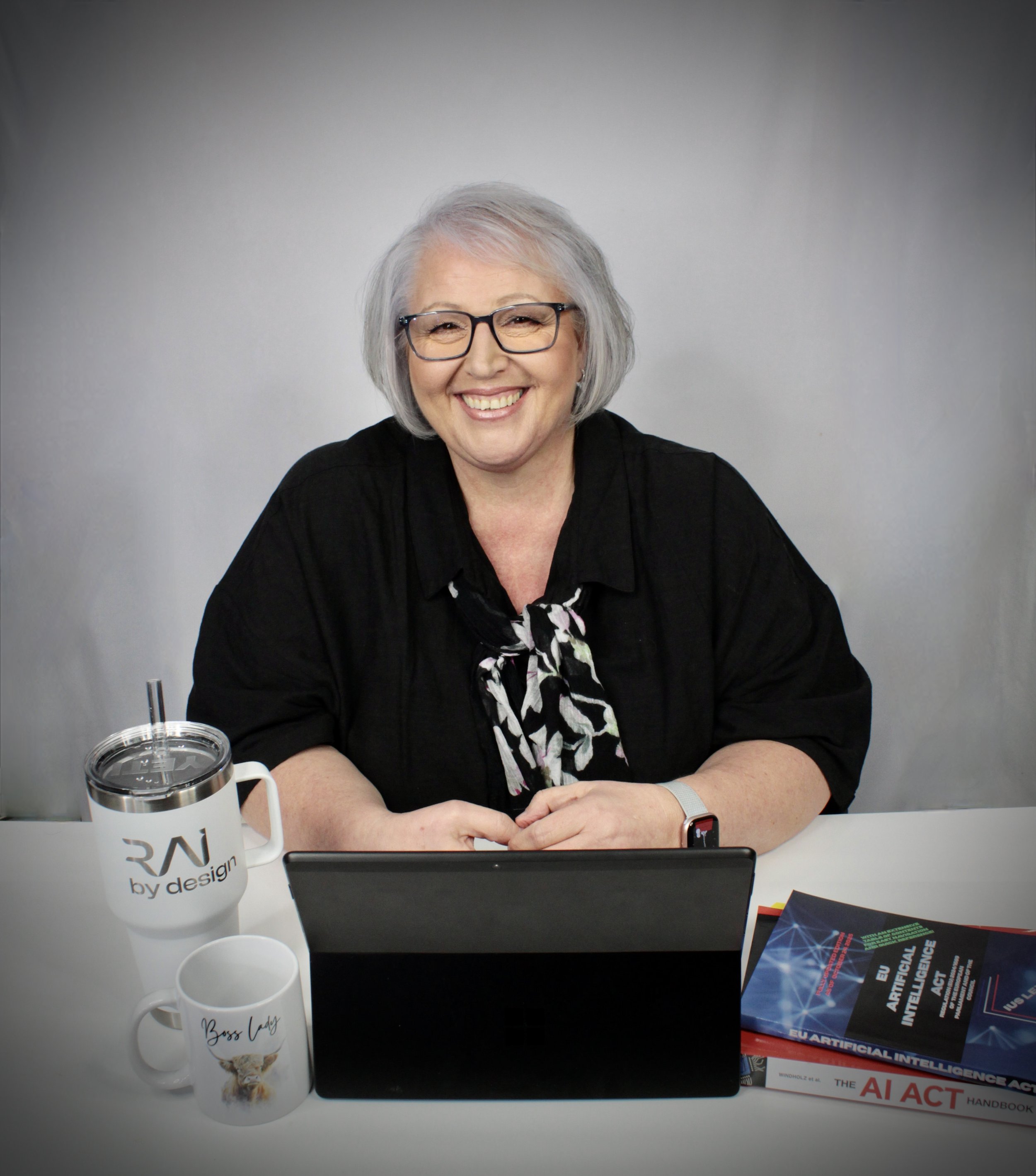 A smiling elderly woman with glasses and grey hair sitting at a white table with a laptop, a mug, a tumbler, and books related to artificial intelligence.