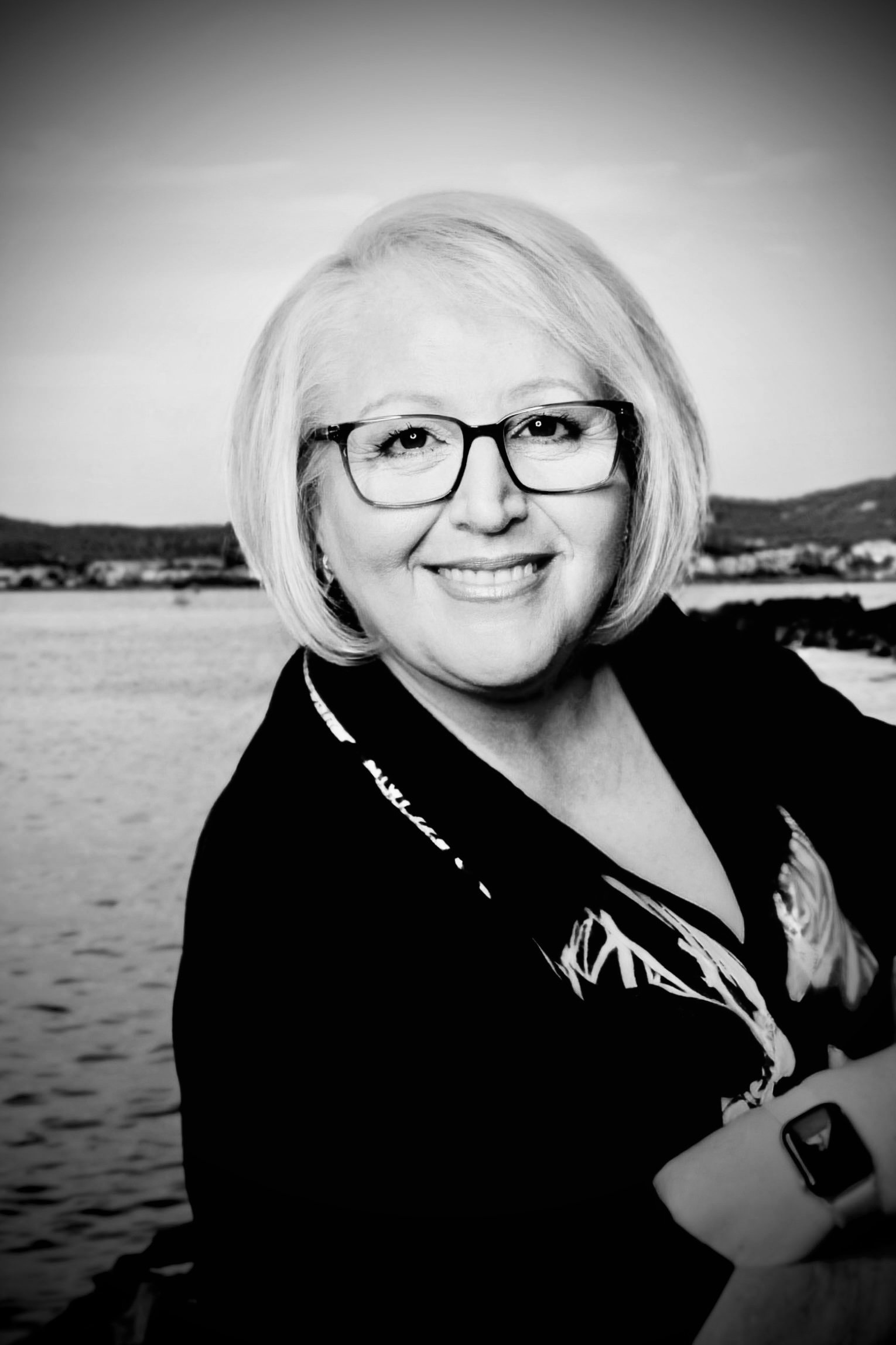 A woman with blond hair, glasses, and a smile, posing outdoors near a body of water, with a distant shoreline in the background.