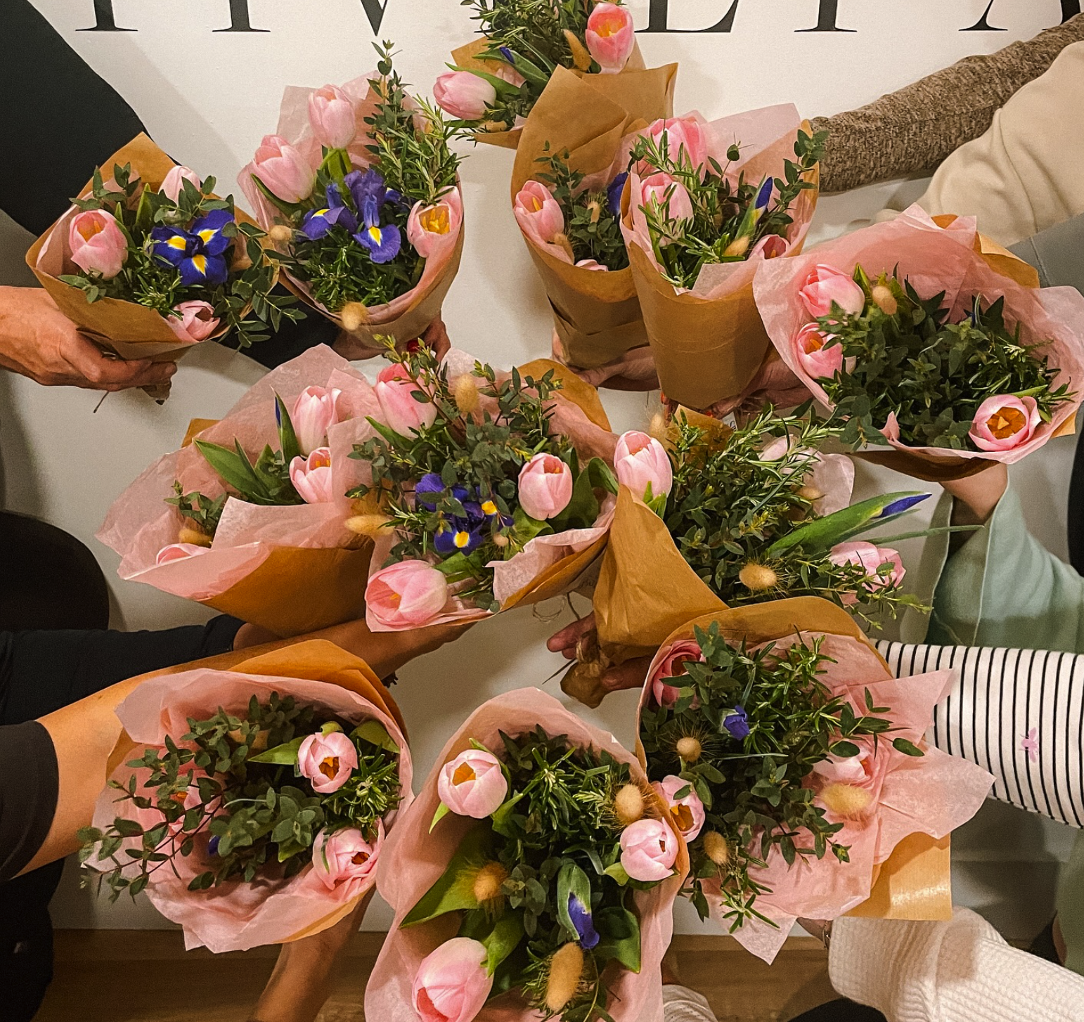 Multiple bouquets of pink tulips and purple irises with green foliage, wrapped in pink and brown paper, being held by several people.