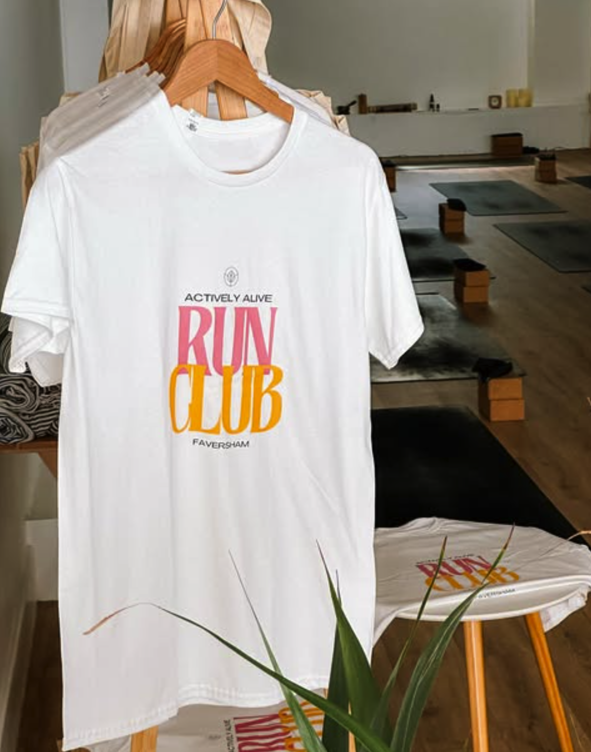 White T-shirt on display with colorful text reading "Actively Alive Run Club Faversham" in pink, orange, and black, hanging on a wooden hanger in a room with yoga mats and wooden blocks in the background.