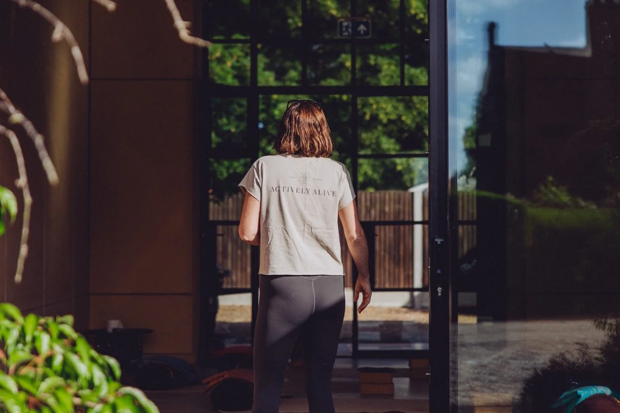 A woman walking into a room through a glass door during daylight, with trees visible outside.