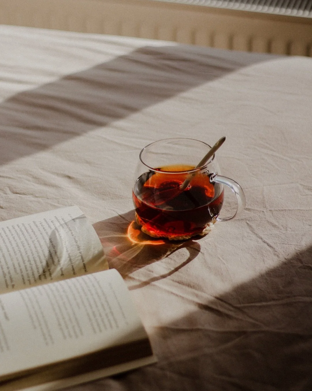 A clear glass cup of hot tea with a spoon inside, placed on a bed with beige bedding, next to an open book, with warm sunlight casting shadows.