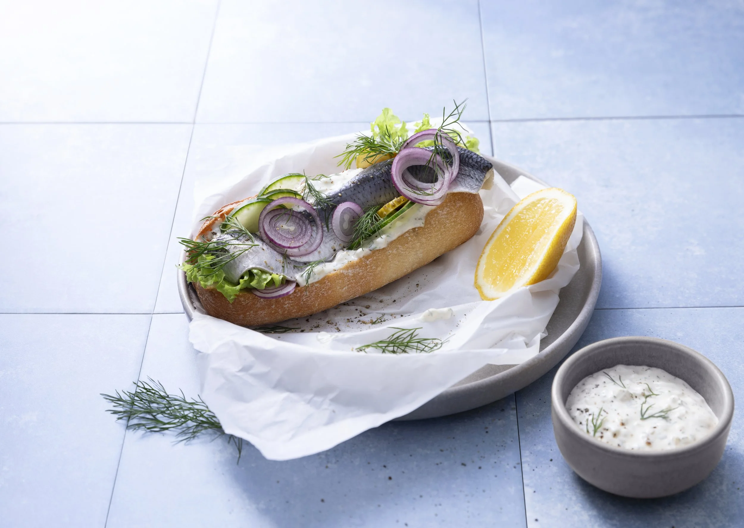Ein Fischbrötchen mit frischen Zutaten, Zitronenscheibe und eine Beilage mit Joghurt-Dip, auf einem blauen Fliesenboden.