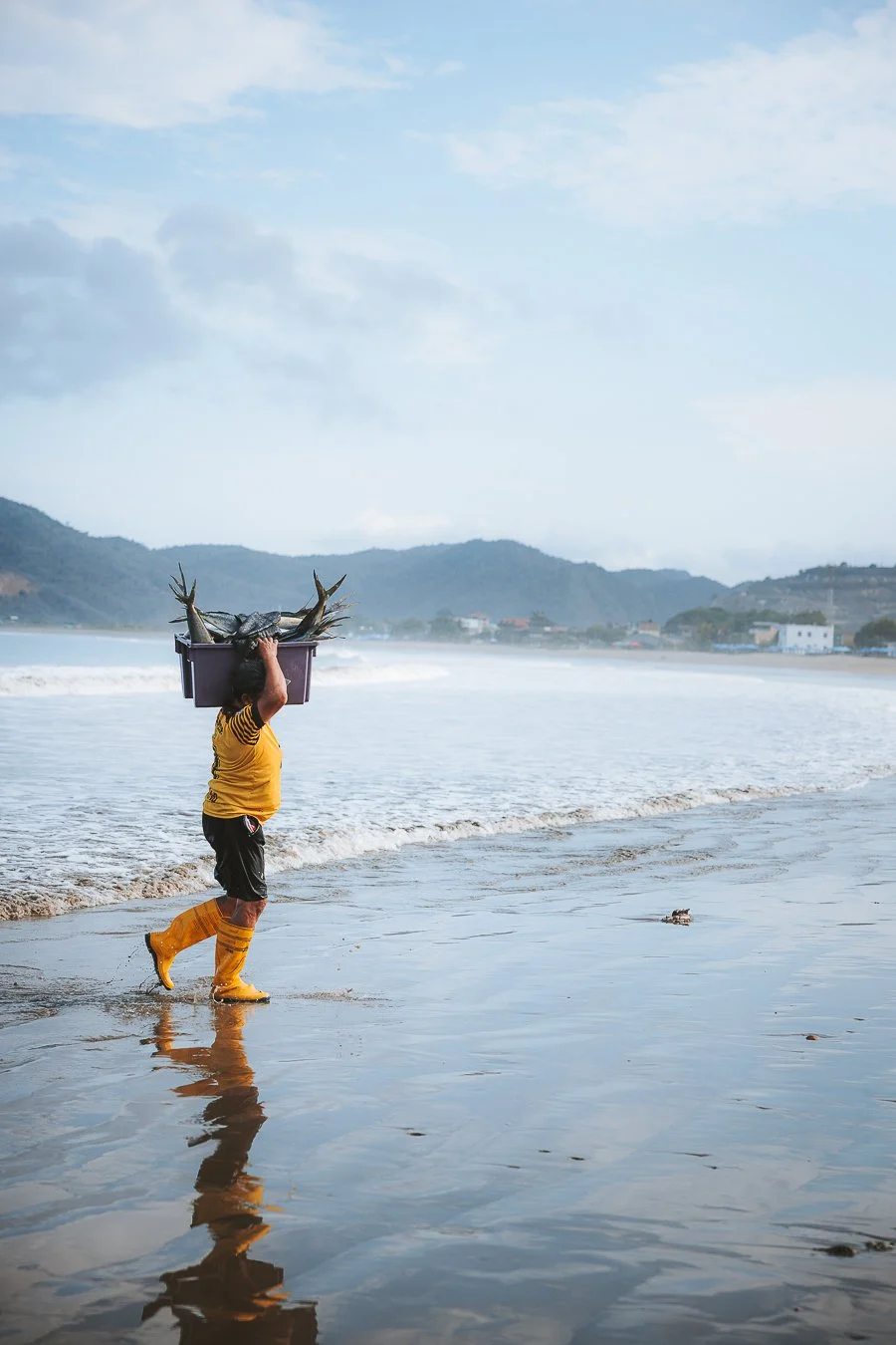 Eine Person, die am Strand im Wasser steht und einen Korb voller Fische trägt.