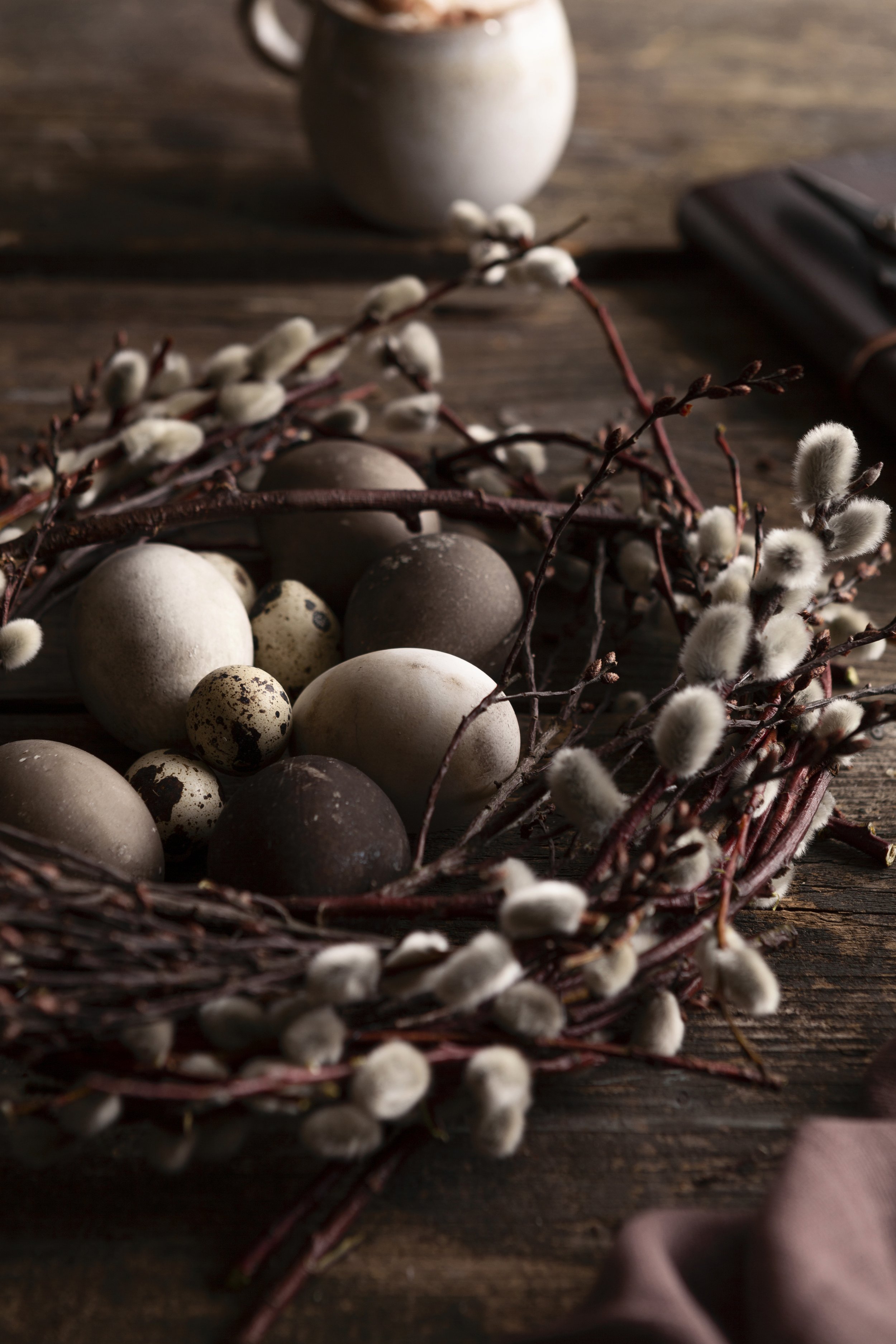Osterkörbchen mit verschiedenen Eiern und Zweigen mit zusammengedrückten Hasennestchen, auf einem Holztisch.