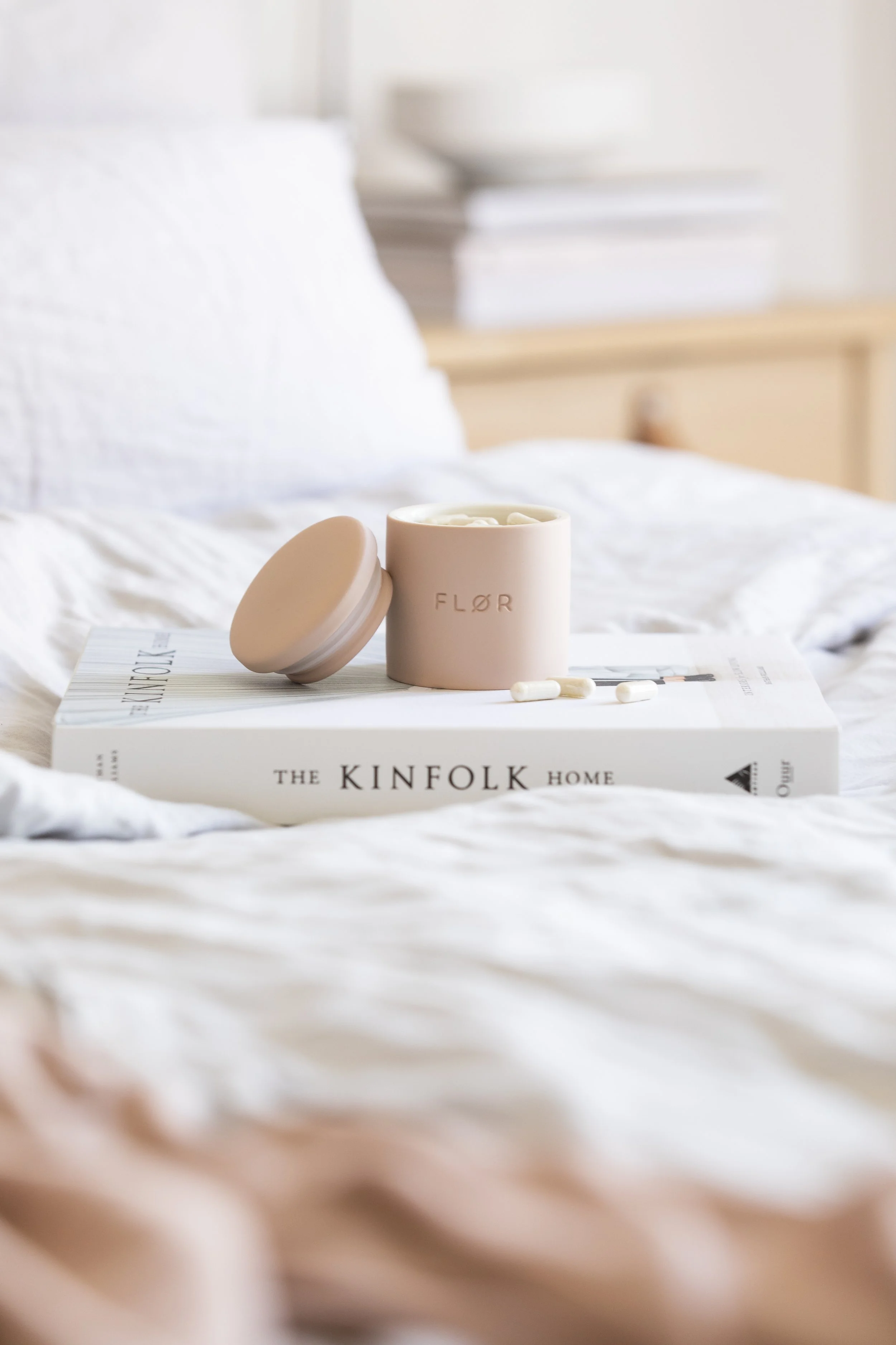 Auf einem Bett liegt ein Buch namens 'The Kinfolk Home' mit einer rosa Kerze und weißen Kapseln darauf.