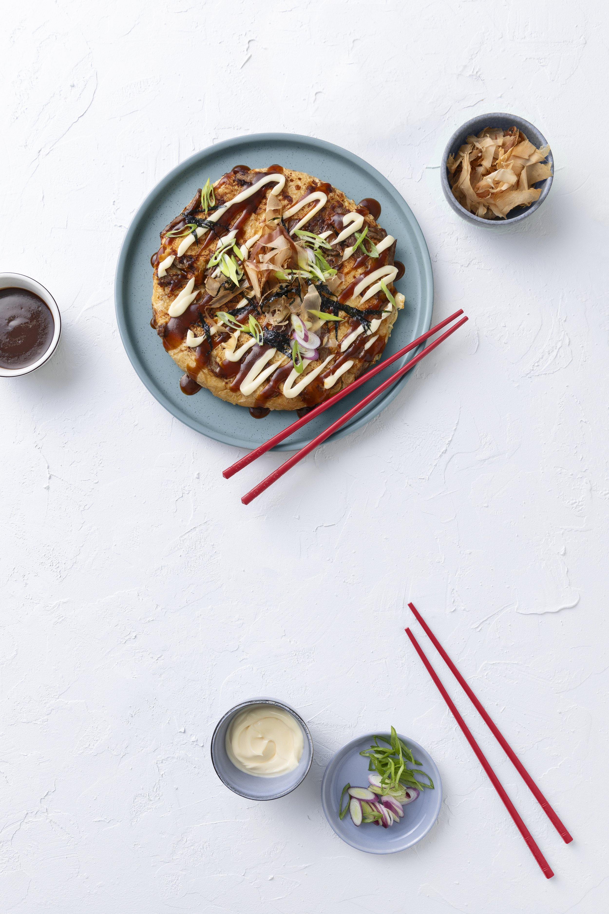 Ein Teller mit japanischer Okonomiyaki, garniert mit Soßen, Frühlingszwiebeln und Bonitoflocken, mit Essstäbchen daneben, auf weißem Hintergrund.