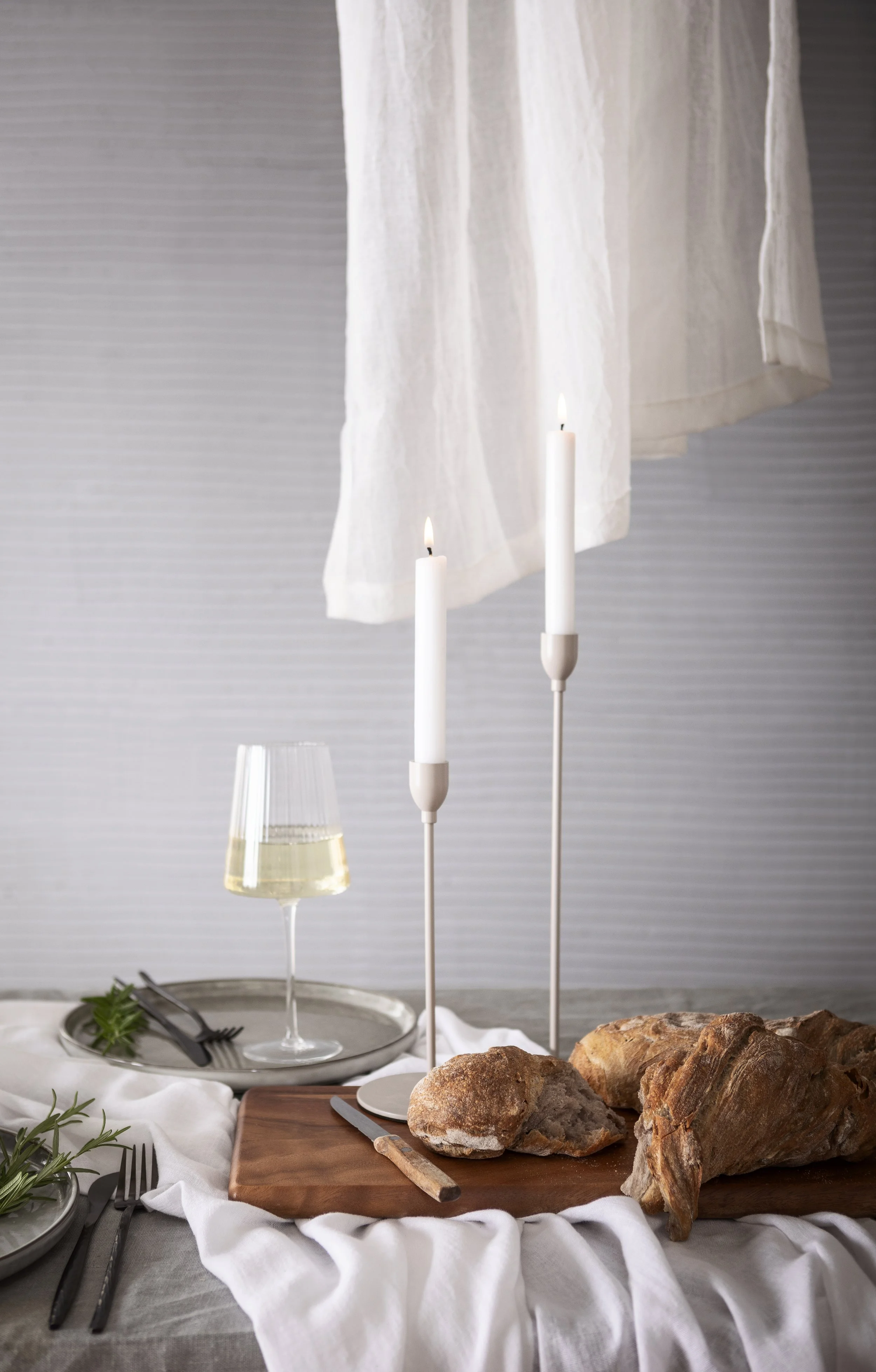 Ein gedeckter Esstisch mit Brot, einem Glas Weißwein, zwei Kerzen in Ständern, Besteck, einem weißen Tuch und einem weißen Vorhang im Hintergrund.