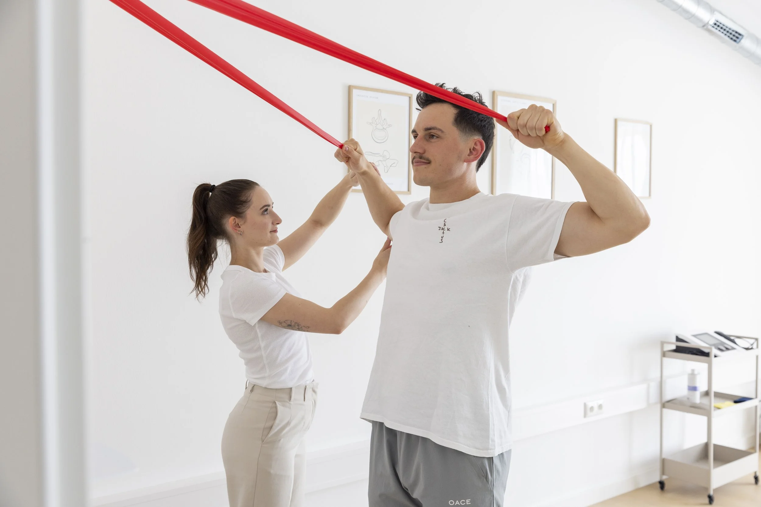 Ein Mann macht eine physische Übung mit einem roten Theraband, während eine Frau ihn dabei anleitet, in einem Physiotherapie- oder Trainingsraum.