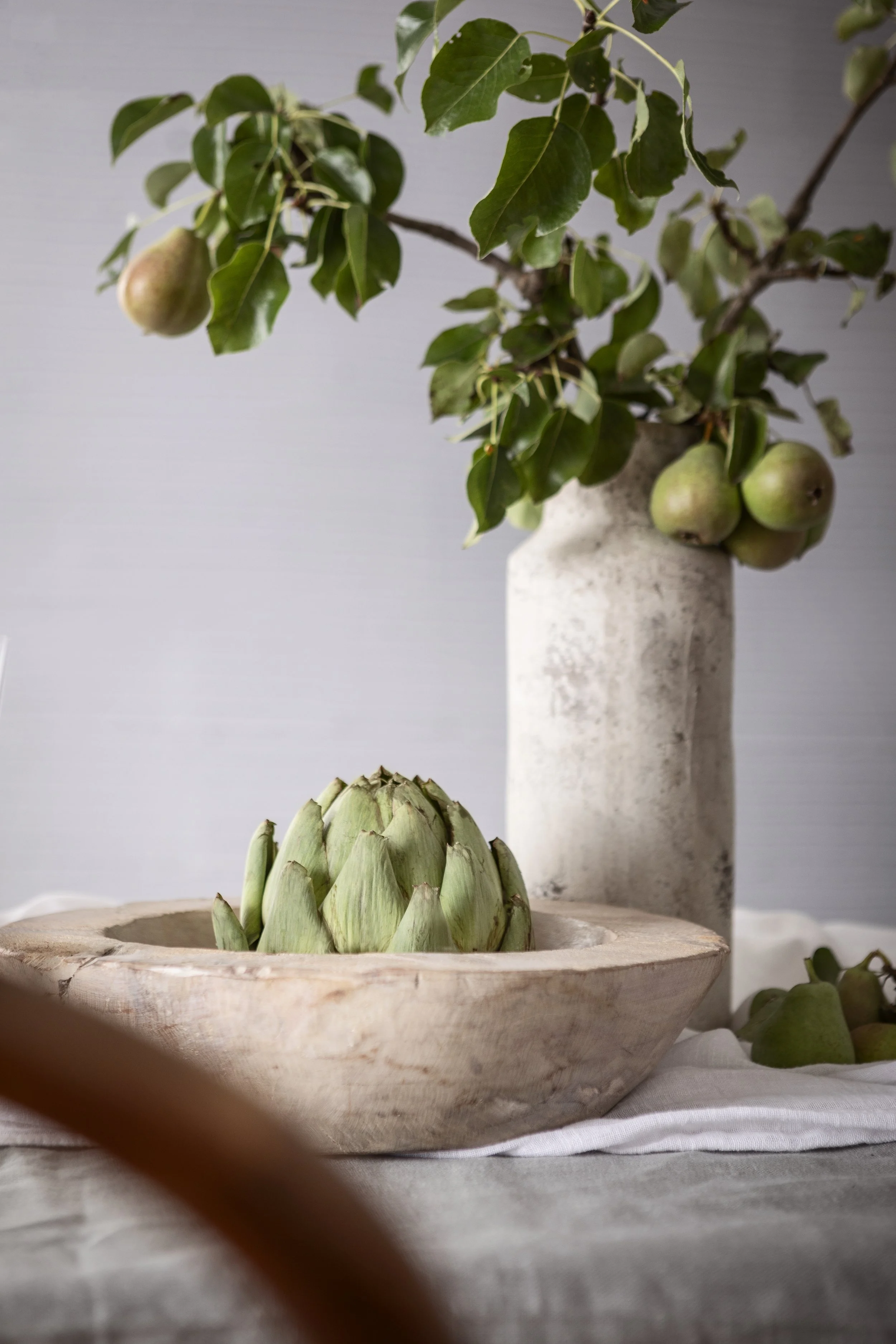 Eine Artischocke in einer Holtschale auf einem Tisch, im Hintergrund ein Blumentopf mit Äpfeln und grünen Blättern, alles in neutralen, natürlichen Farben.