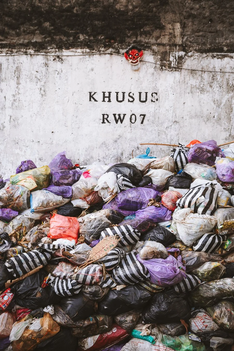 Haufen von Müllsäcken und Plastikabfällen vor einer weißen Wand mit schwarzen Buchstaben, die Khusus RW07 schreiben, und eine kleine rote Maskenfigur oben an der Wand.