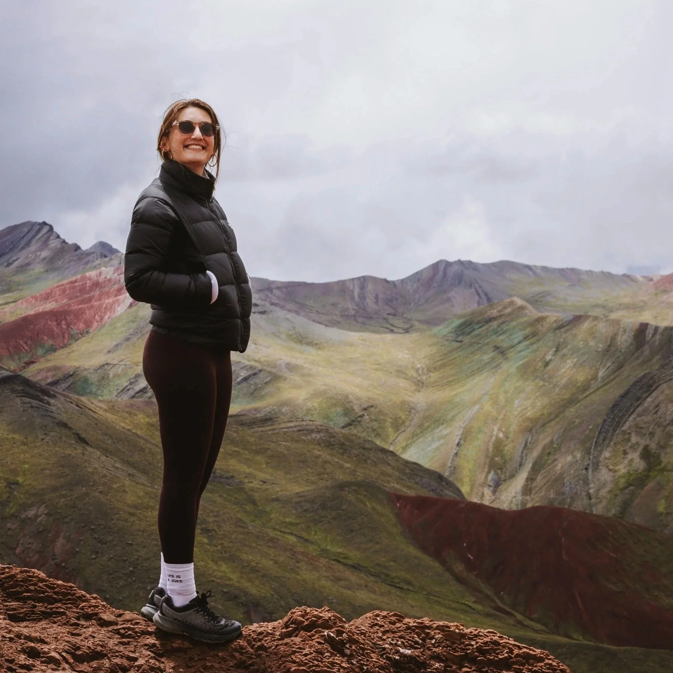 Eine lachende Frau mit dunkler Jacke, Sonnenbrille, schwarzen Leggings, weißen Socken und Sportschuhen steht auf rotem Felsen vor einer bunten, bergigen Landschaft bei bewölktem Himmel.