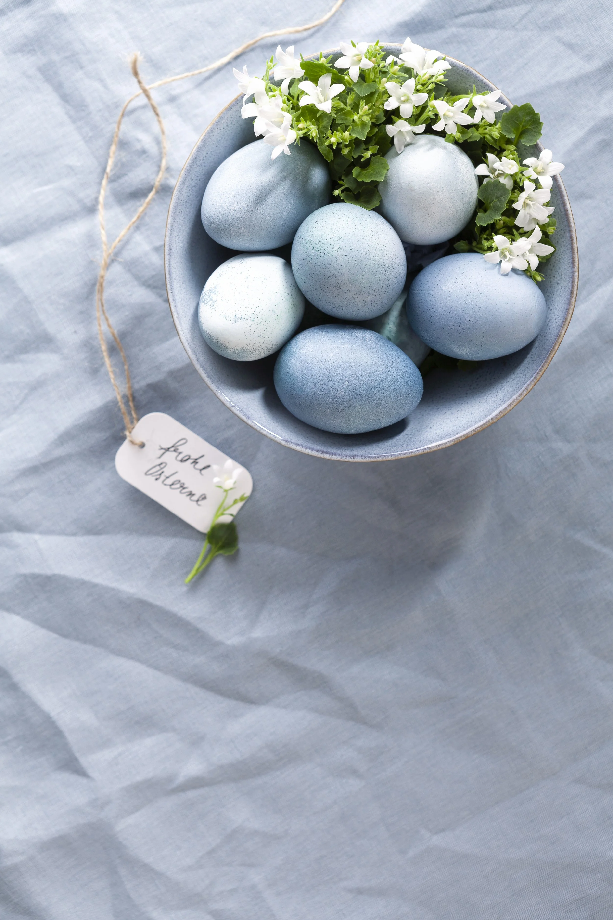 Osterkörbchen mit blauen Ostereiern und weißen Frühlingsblumen, darunter ein kleiner Anhänger mit der Aufschrift 'Frohe Ostern'.