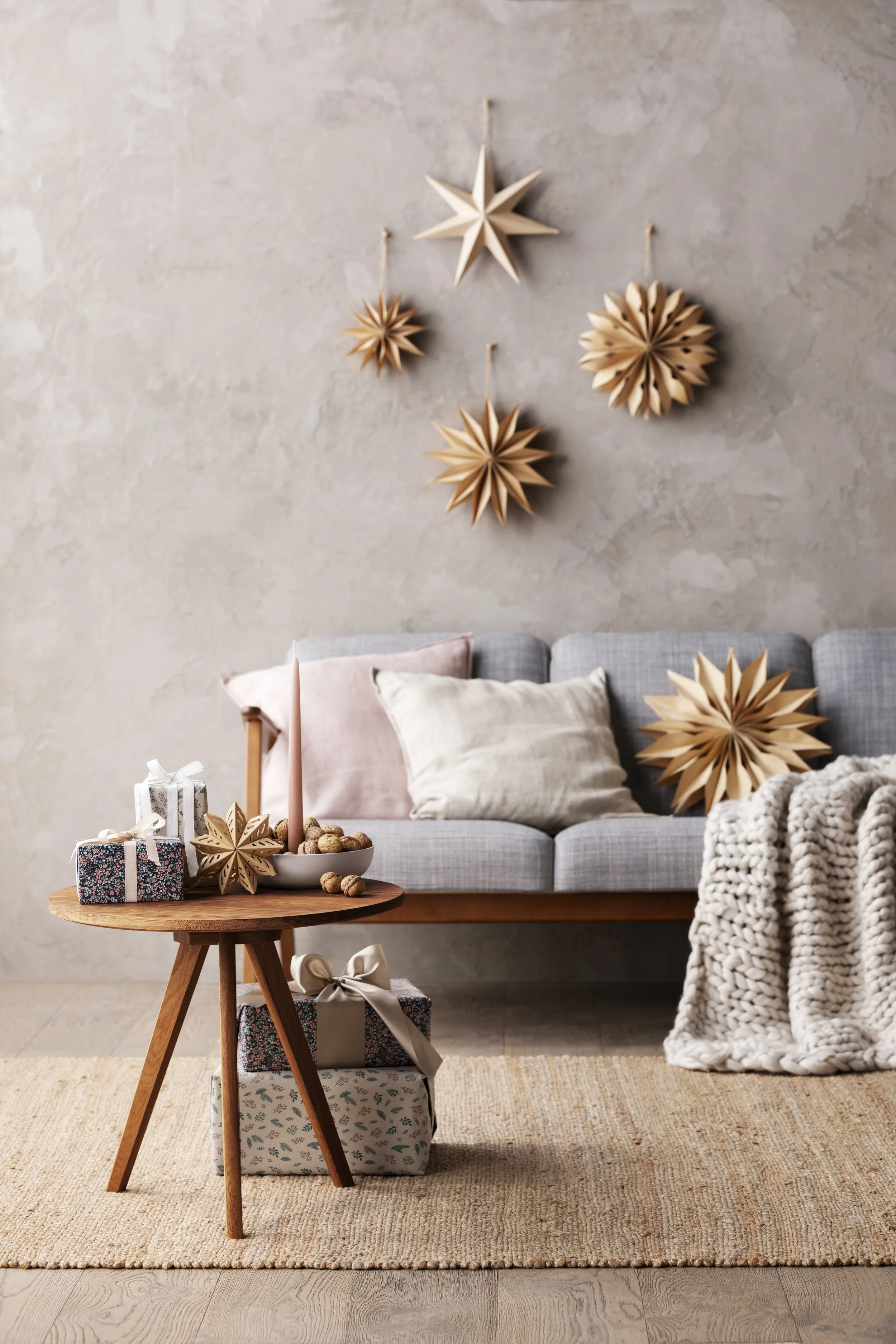 Gemütliches Wohnzimmer mit Weihnachtsdekoration, bestehend aus geschenkverpackungen, Weihnachtssterne aus Holz an der Wand und einer grauen Couch mit Kissen und einer Strickdecke.