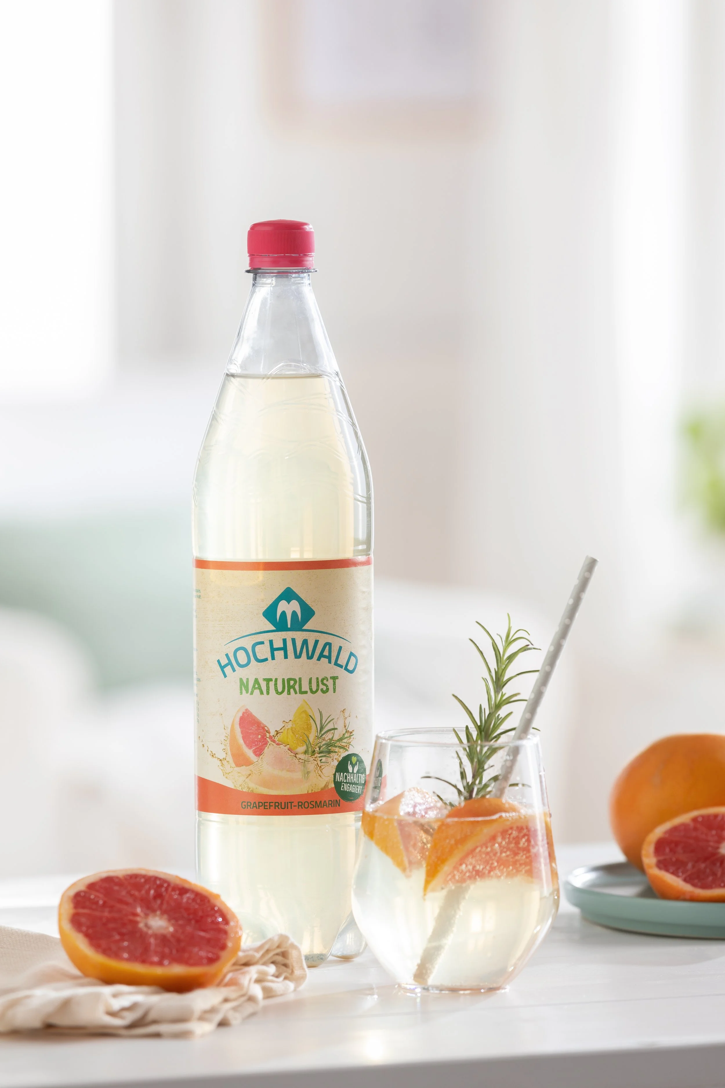 Eine Flasche Grapefruit-Rosmarin-Erfrischungsgetränk mit eine Glas, darin sind Grapefruit- und Rosmarinzweige, auf einem weißen Tisch mit geschnittener Grapefruit und Orangen. Im Hintergrund ist ein unscharfer Raum.