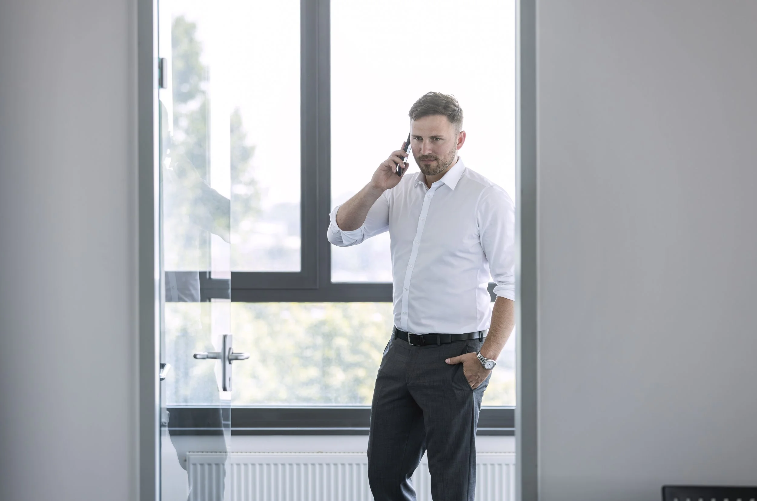 Ein Mann in Business-Kleidung steht vor einem Fenster und telefoniert.
