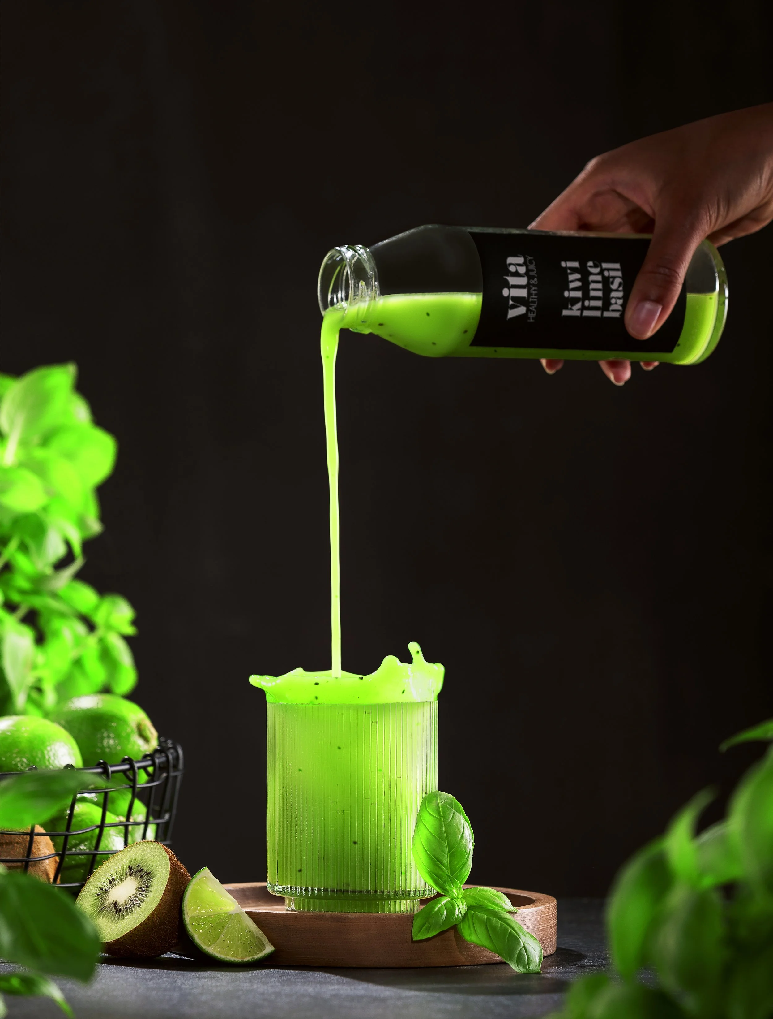 Hand, die einen grünen grünen Saft namens "Kivi Basil" aus einer Flasche in ein Glas gießt, umgeben von frischen Basilikumblättern, Kiwi, Limetten und grünen Lemons. Hintergrund ist dunkel.
