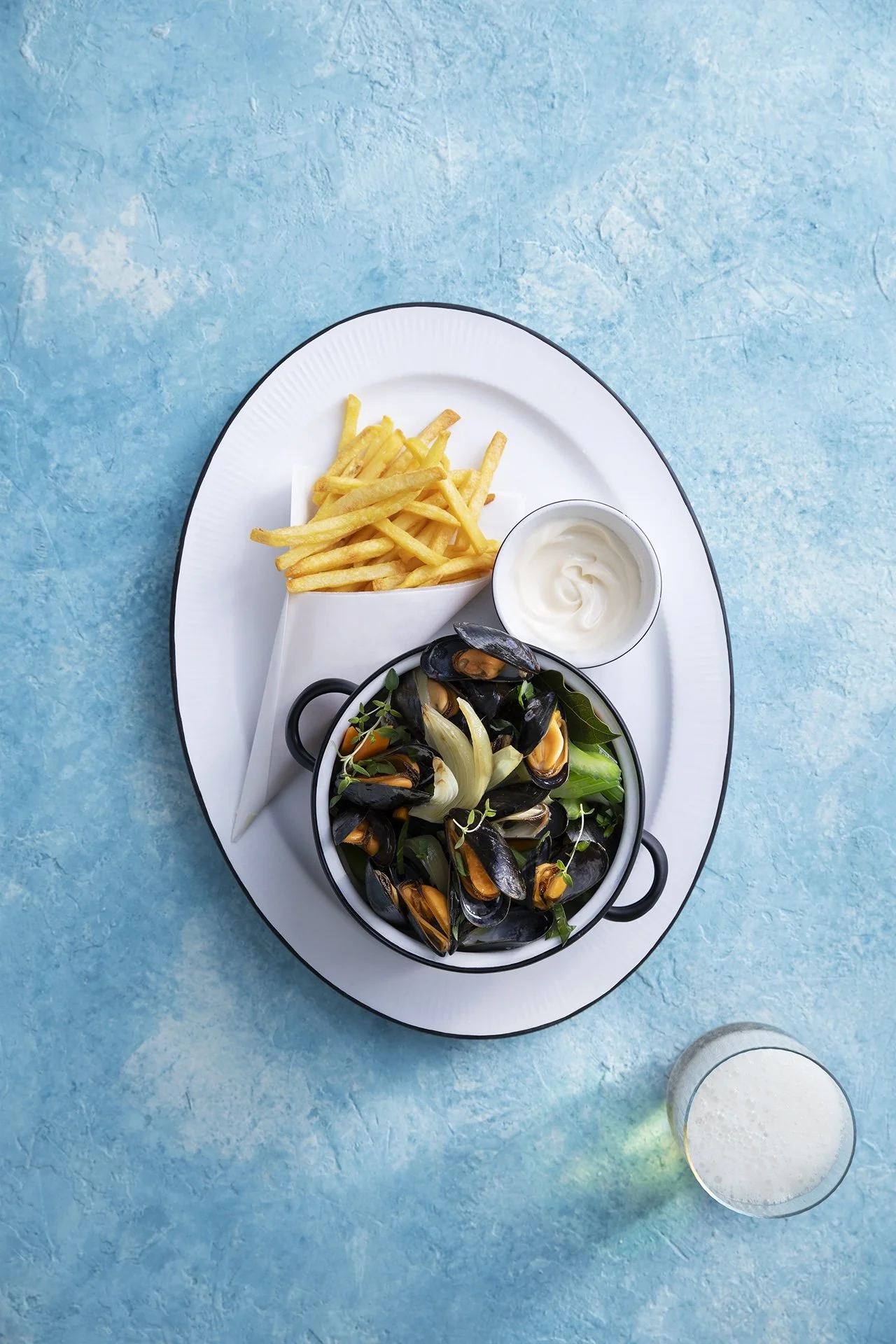 Auf einem weißen ovalen Teller befinden sich Pommes, eine Schüssel mit Joghurt, eine kleine Pfanne mit Miesmuscheln und Gemüse, sowie ein Glas mit Milch oder einem Getränk. Der Teller liegt auf einem blauen Tisch.