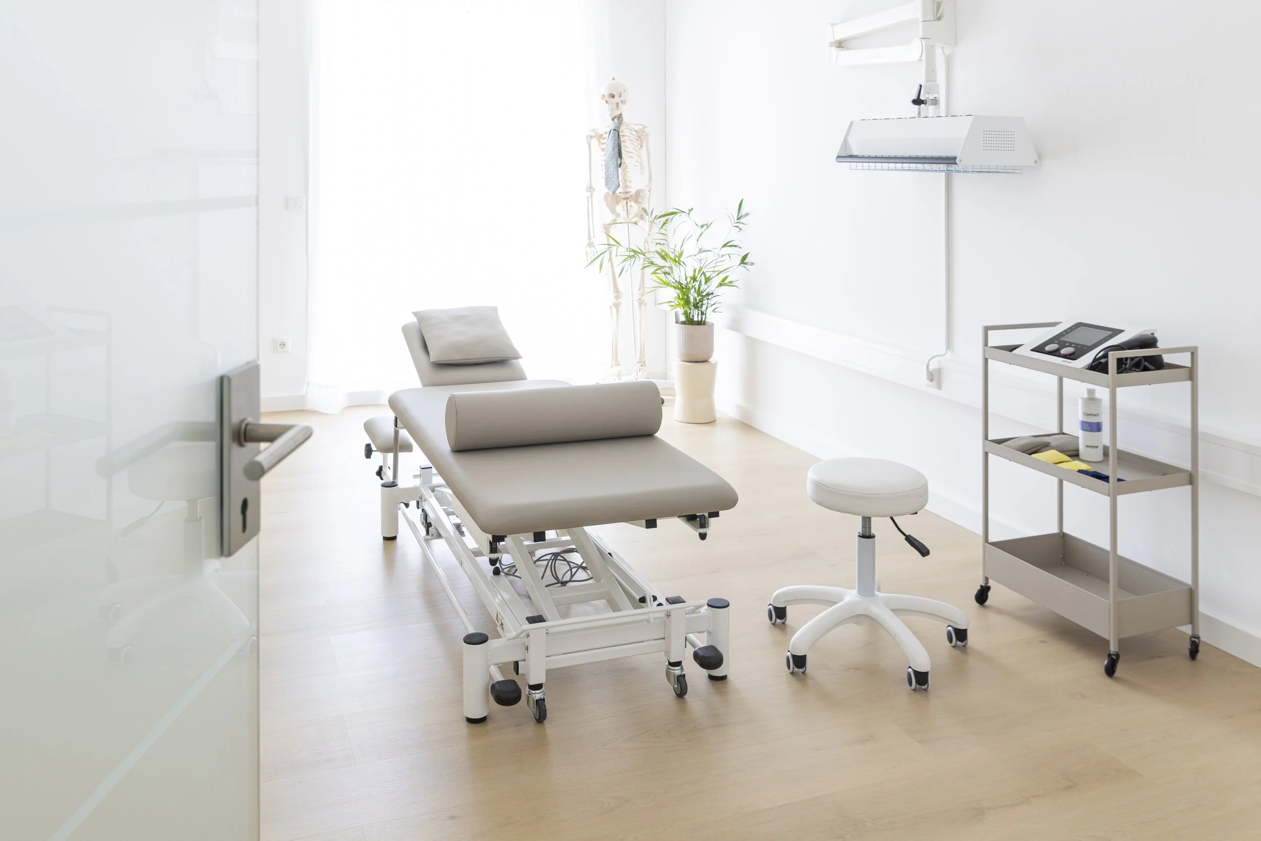 Ein leeres medizinisches Behandlungszimmer mit einem Untersuchungstisch, Stuhl, Pflanze, Modell des menschlichen Skeletts, medizinischen Geräten auf einem Wagen und einer weißen Wand.