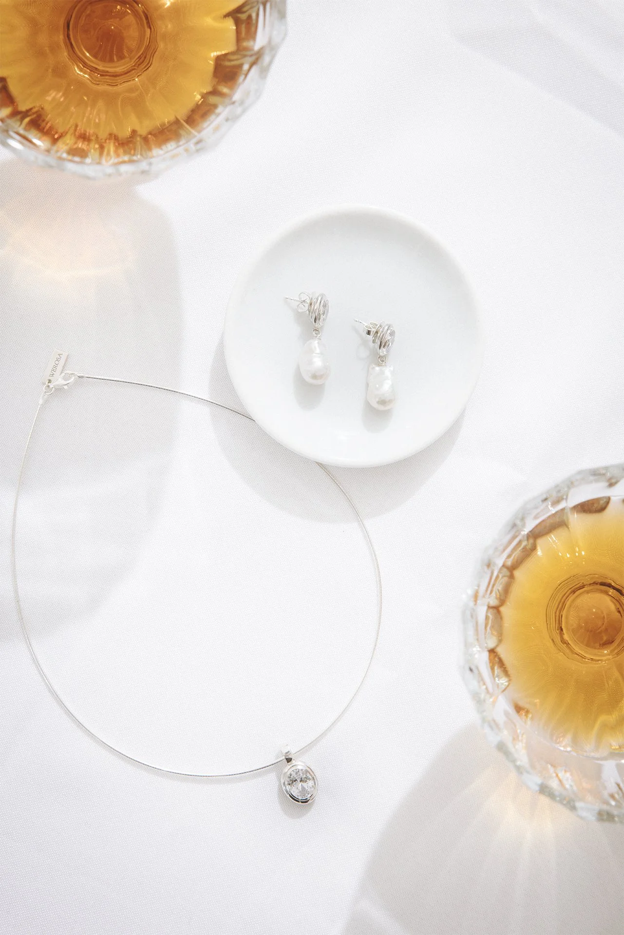 Eine weiße Tafel mit Schmuck, darunter eine Perlenkette, eine Edelsteinkette und Ohrringe mit Perlen, umgeben von zwei Kerzen in Kristallvasen.