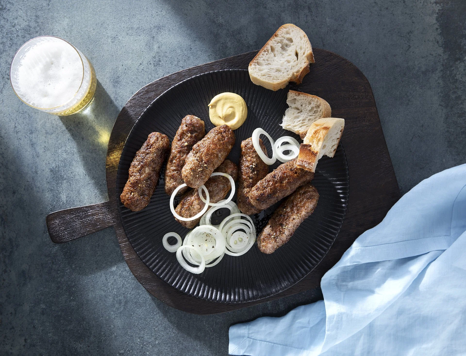 Gericht mit Bratwürsten, Zwiebelringen, Senf und Brot auf einem schwarzen Teller, daneben ein Glas Bier.