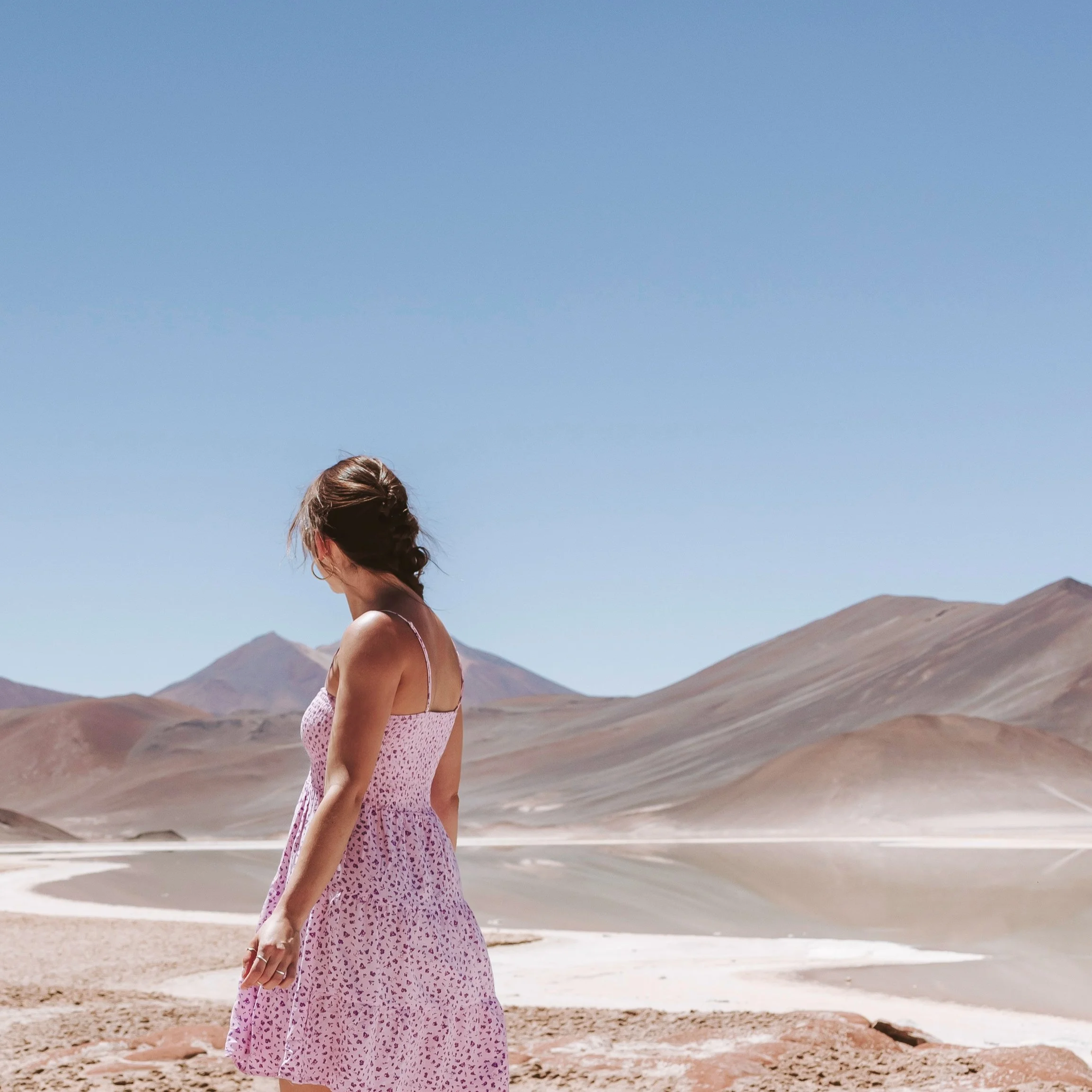 Eine Frau in einer rosa, gemusterten Sommerkleidung steht in einer Wüstenlandschaft mit Bergen im Hintergrund, under einem klaren blauen Himmel.