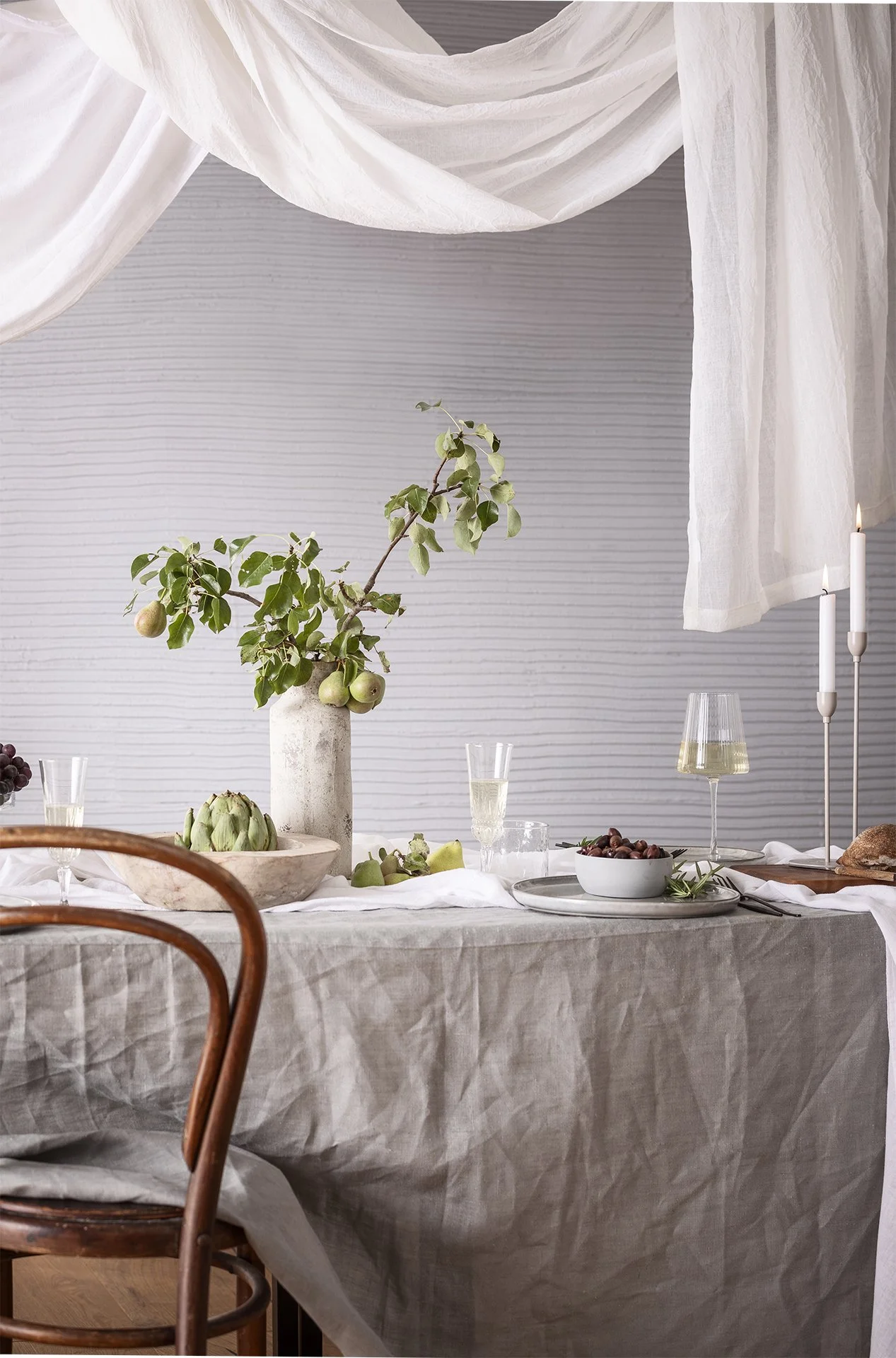 Ein gedeckter Esstisch mit einer weißen Tischdecke, einem Weinblatt in einer Vase, einer Schale mit Artischocken, einem Glas Sekt, einer weißen Schüssel mit Trauben, Kerzen und Weißtuch-Dekorationen.