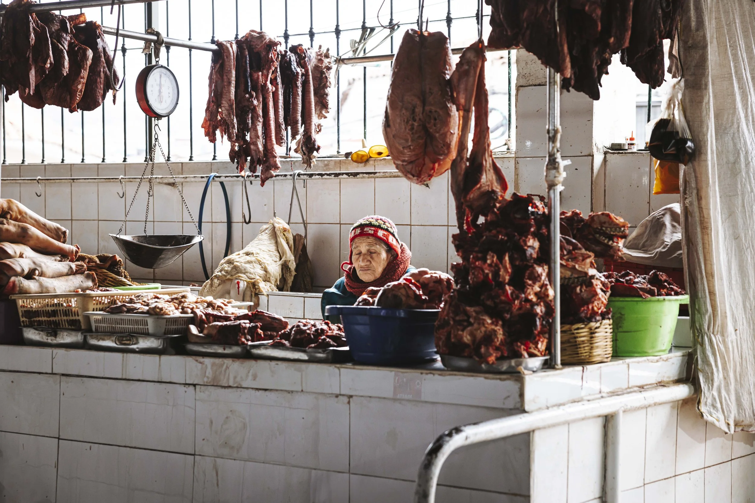 Eine Marktfrau verkauft Fleisch in einem Marktladen, mit an Haken gehängtem frischem Fleisch und einigen Fleischstücken auf dem Tresen.