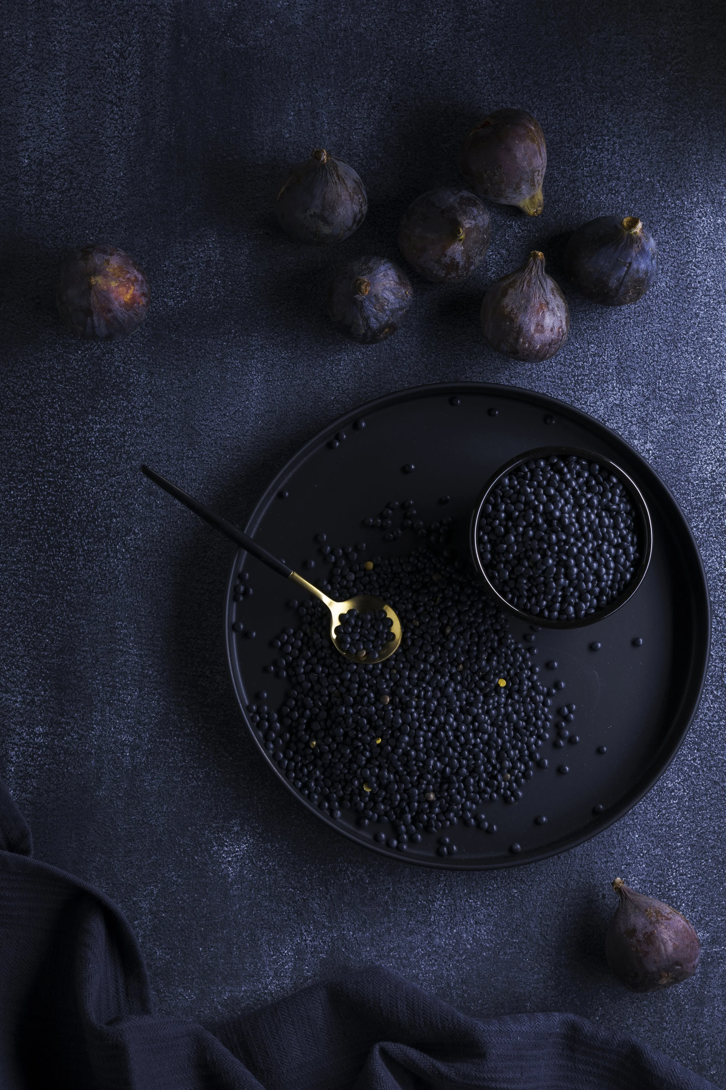 Schwarze Johannisbeerbeeren auf einem schwarzen Teller, einige in einer kleinen Schale, sowie frische Feigen auf einer dunklen Oberfläche.
