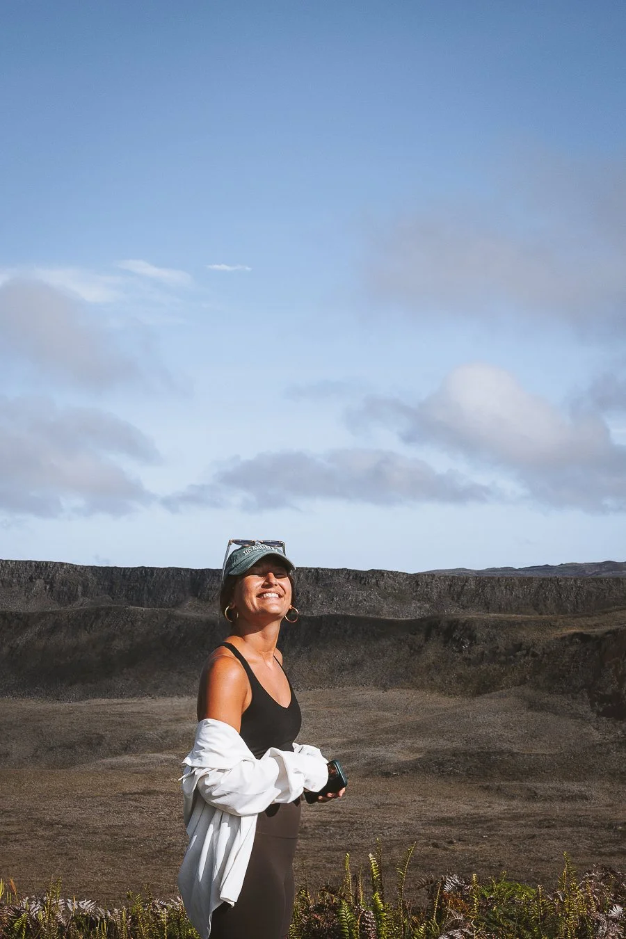 Eine junge Frau steht in einer vulkanischen Landschaft, lächelt und blickt in den Himmel, trägt eine schwarze Top, eine weiße Jacke um die Taille und einen cap, hält ein Smartphone in der Hand.
