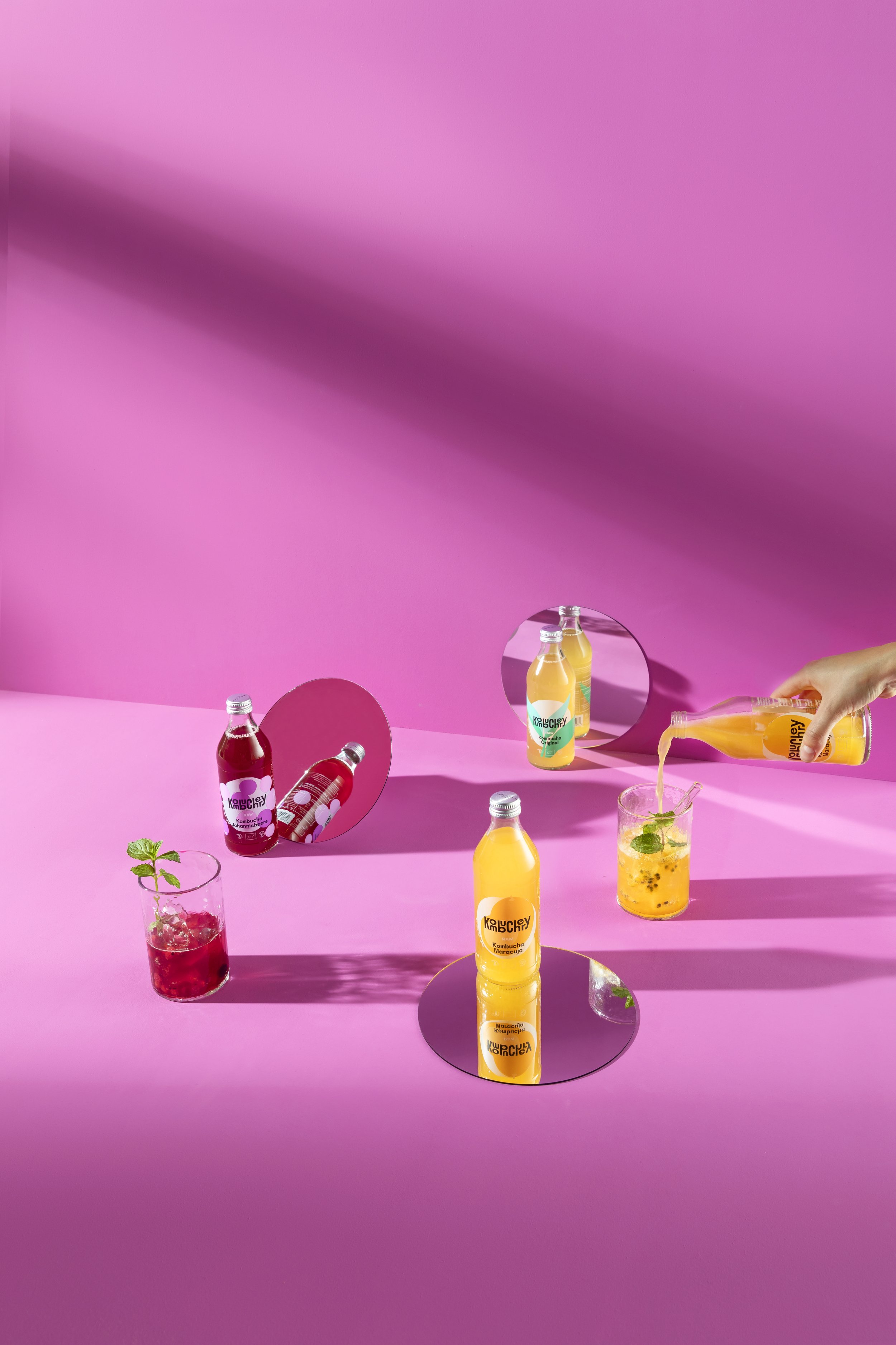 Fünf Flaschen mit bunten Getränken, ein Glas mit Getränk und Minzblättern, sowie eine Person, die eine Flasche eingießt, auf einem pinken Hintergrund, mit Spiegeln. 