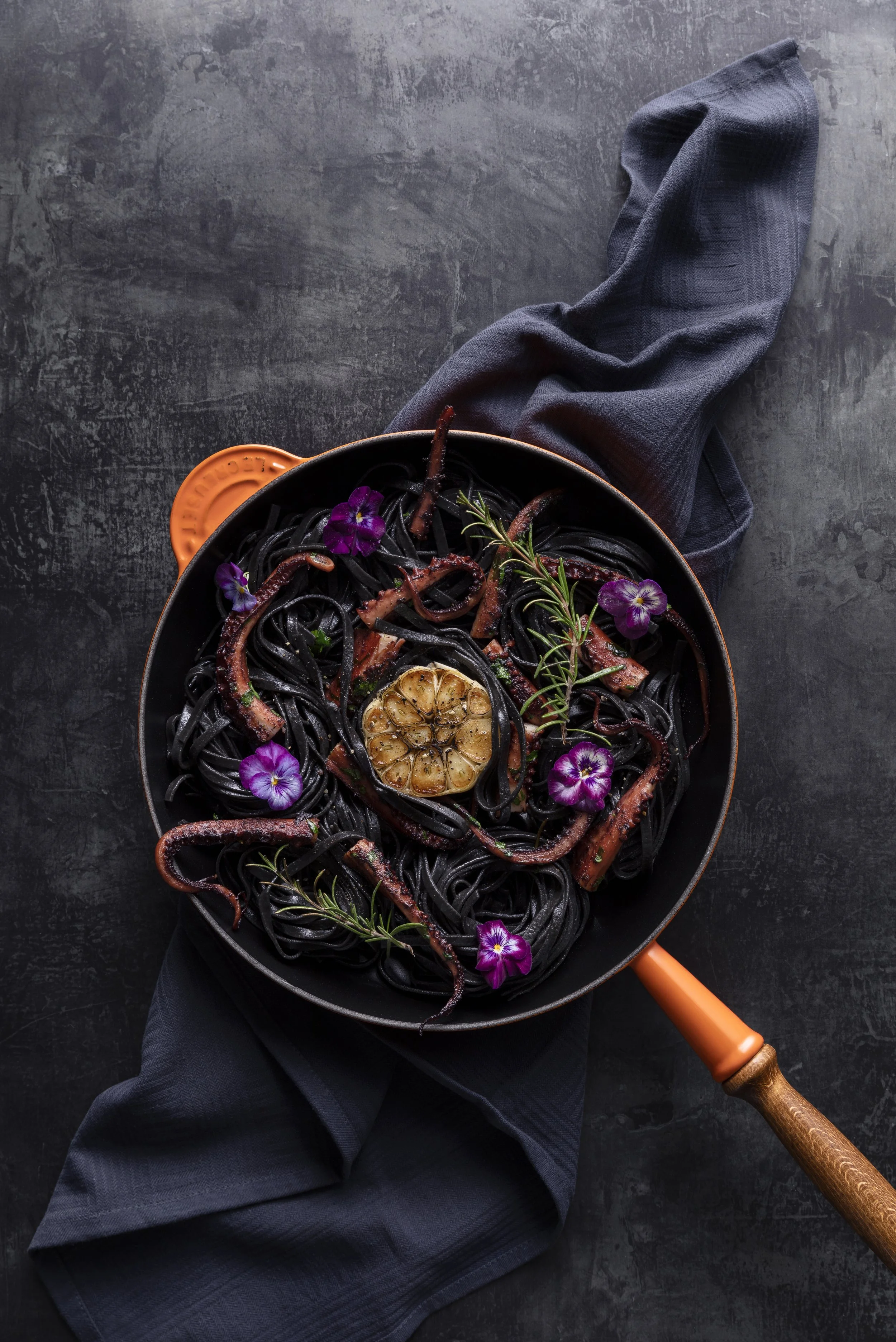 Schwarze Spaghetti mit gebratenen Tintenfischen, Zitronenscheiben, Rosmarin und Veilchenblumen in einer schwarzen Pfanne auf einem dunklen Hintergrund