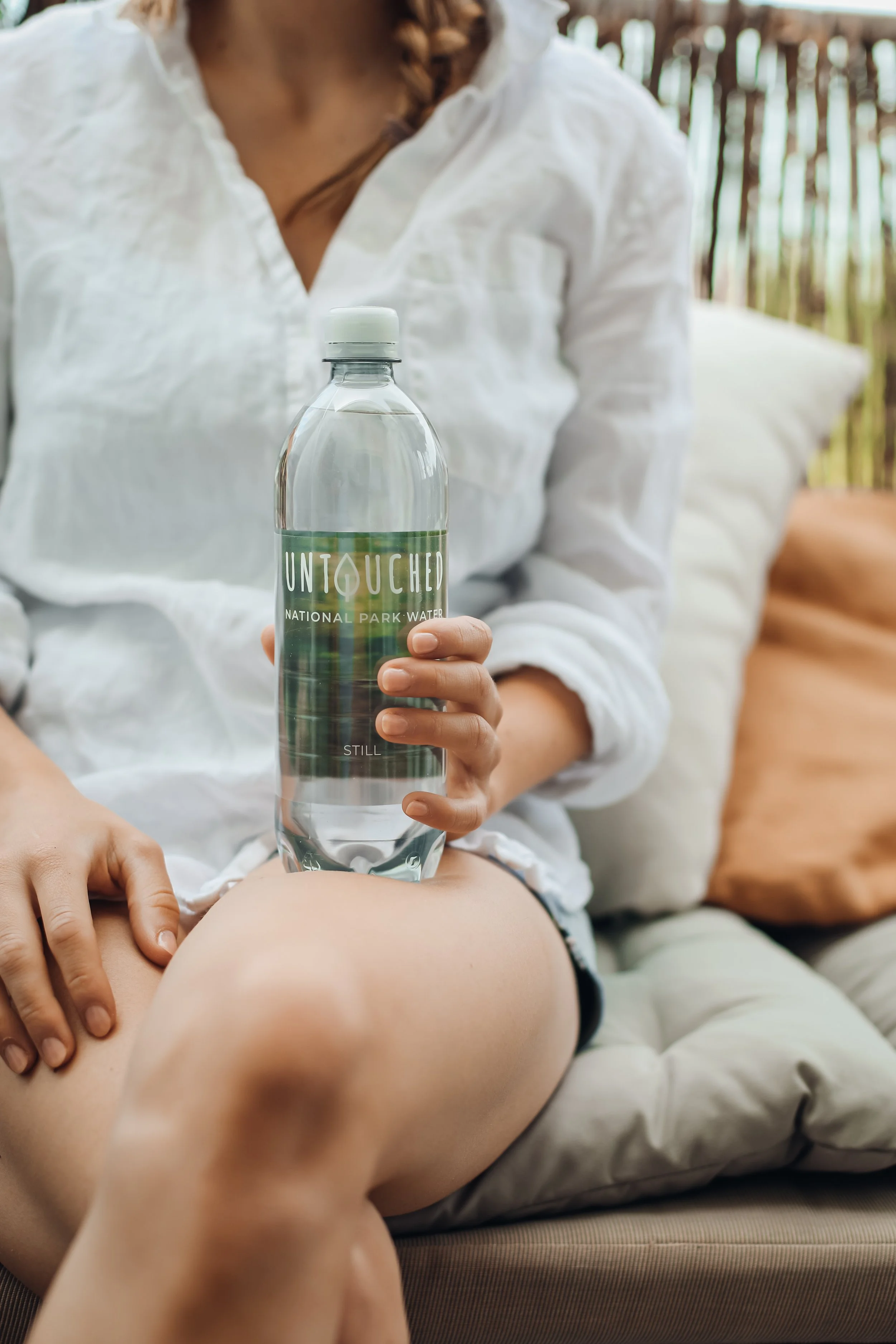 Eine Person in einem weißen Hemd sitzt auf einem Sofa und hält eine Flasche mit Wasser. Die Flasche hat ein Label mit dem Wort "UNTOUCHED" und zeigt ein Naturbild. Die Person hat braune Haare zu Zöpfen und sitzt im Freien vor einer Holzwand.