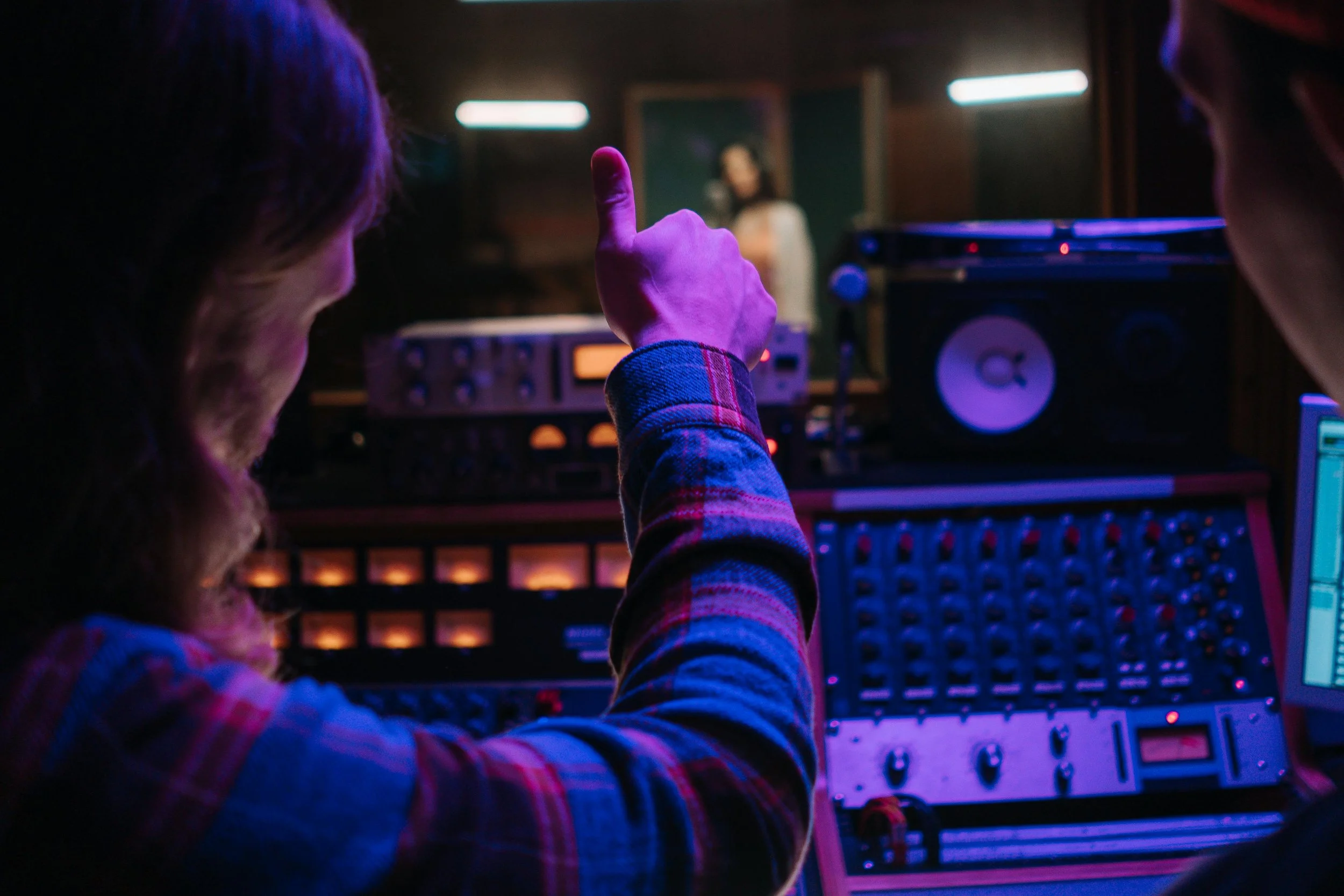 A person in a plaid shirt making a peace sign with their hand in front of music equipment in a dimly lit room, with a woman on a screen in the background.