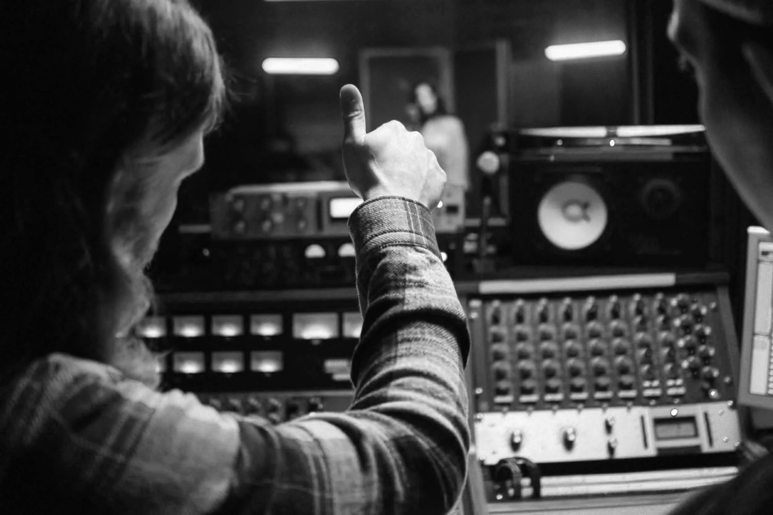 A person in a plaid shirt making a peace sign with their hand in front of music equipment in a dimly lit room, with a woman on a screen in the background.