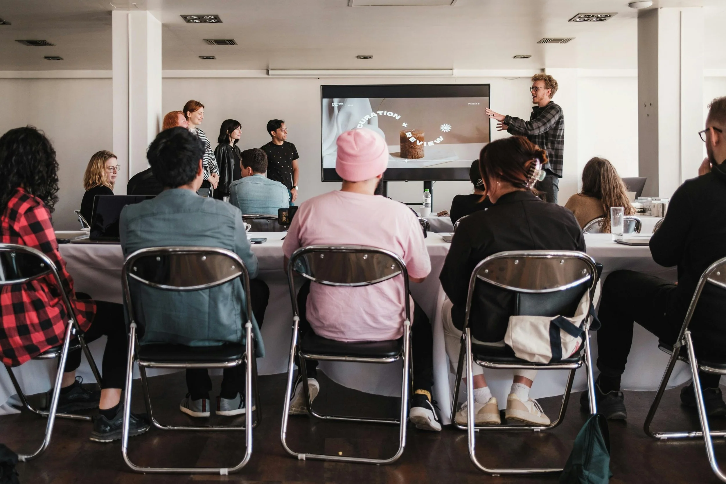 A group of people attending a presentation or workshop in a conference room. The presenter is standing next to a large screen displaying a slide with the words 'Curration + Review' and an image of a cake. Attendees are seated around tables, listening to the speaker.