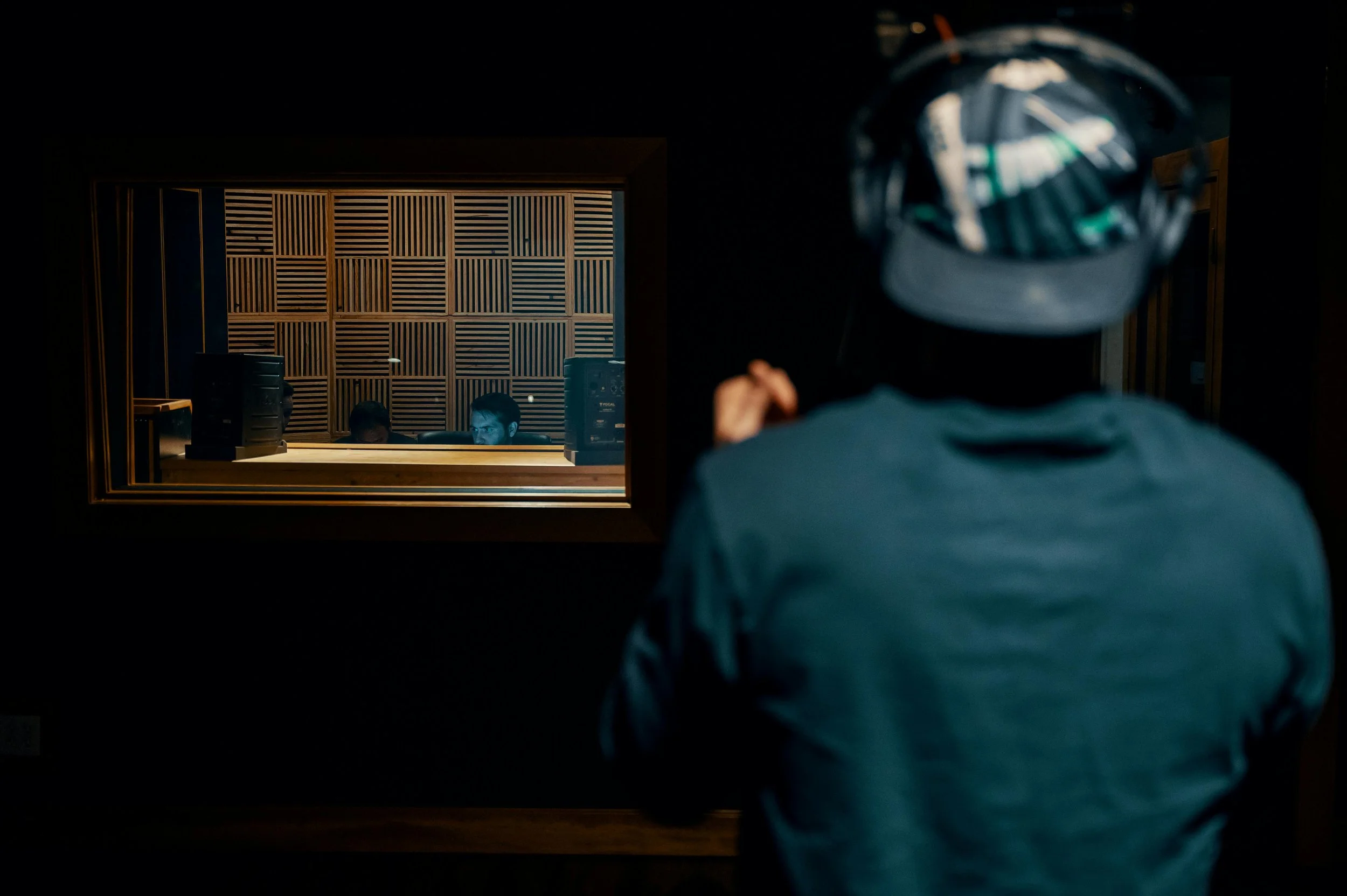 A person wearing a cap and jacket faces away from the camera, looking into a recording studio window where two people are working at a mixing console.