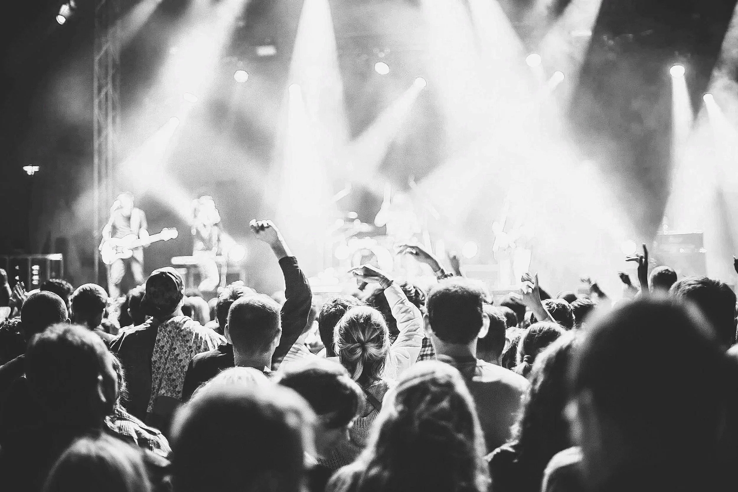 A large crowd of people attending a live concert with a band performing on stage, illuminated by bright stage lights.