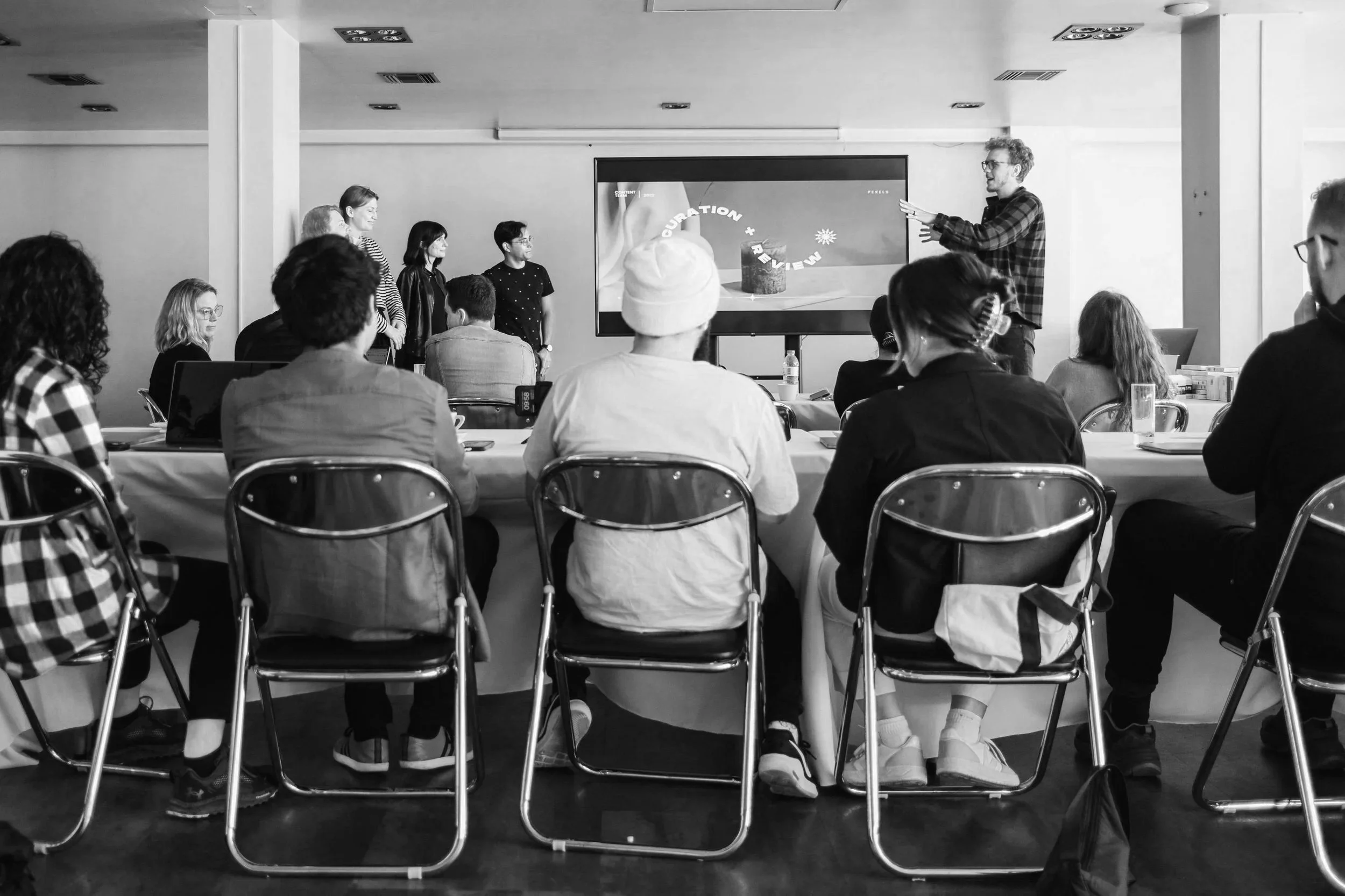 A group of people attending a presentation or workshop in a conference room. The presenter is standing next to a large screen displaying a slide with the words 'Curration + Review' and an image of a cake. Attendees are seated around tables, listening to the speaker.