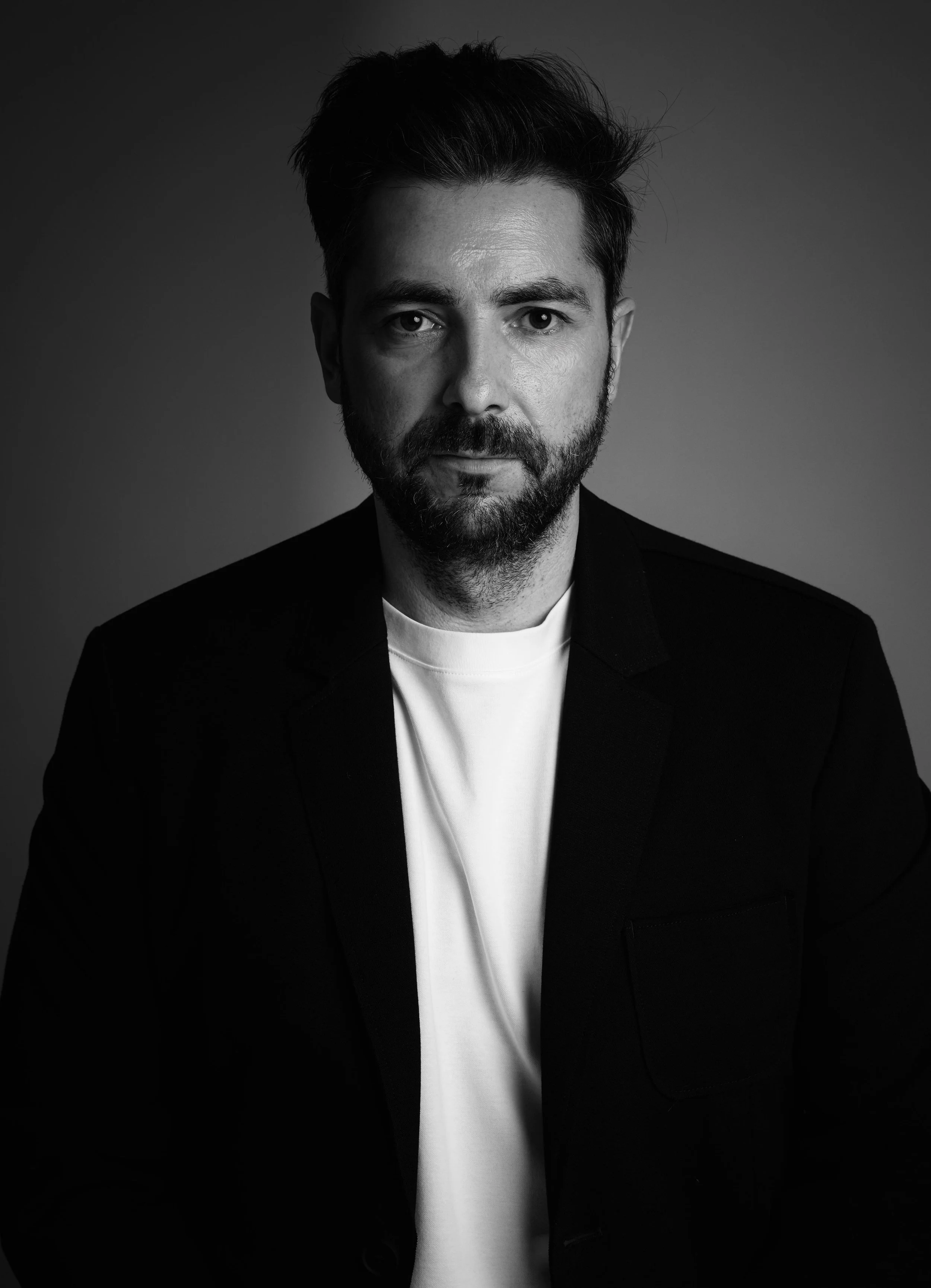 Black and white portrait of a man with dark hair and a beard, wearing a dark blazer and a white shirt, looking directly at the camera.